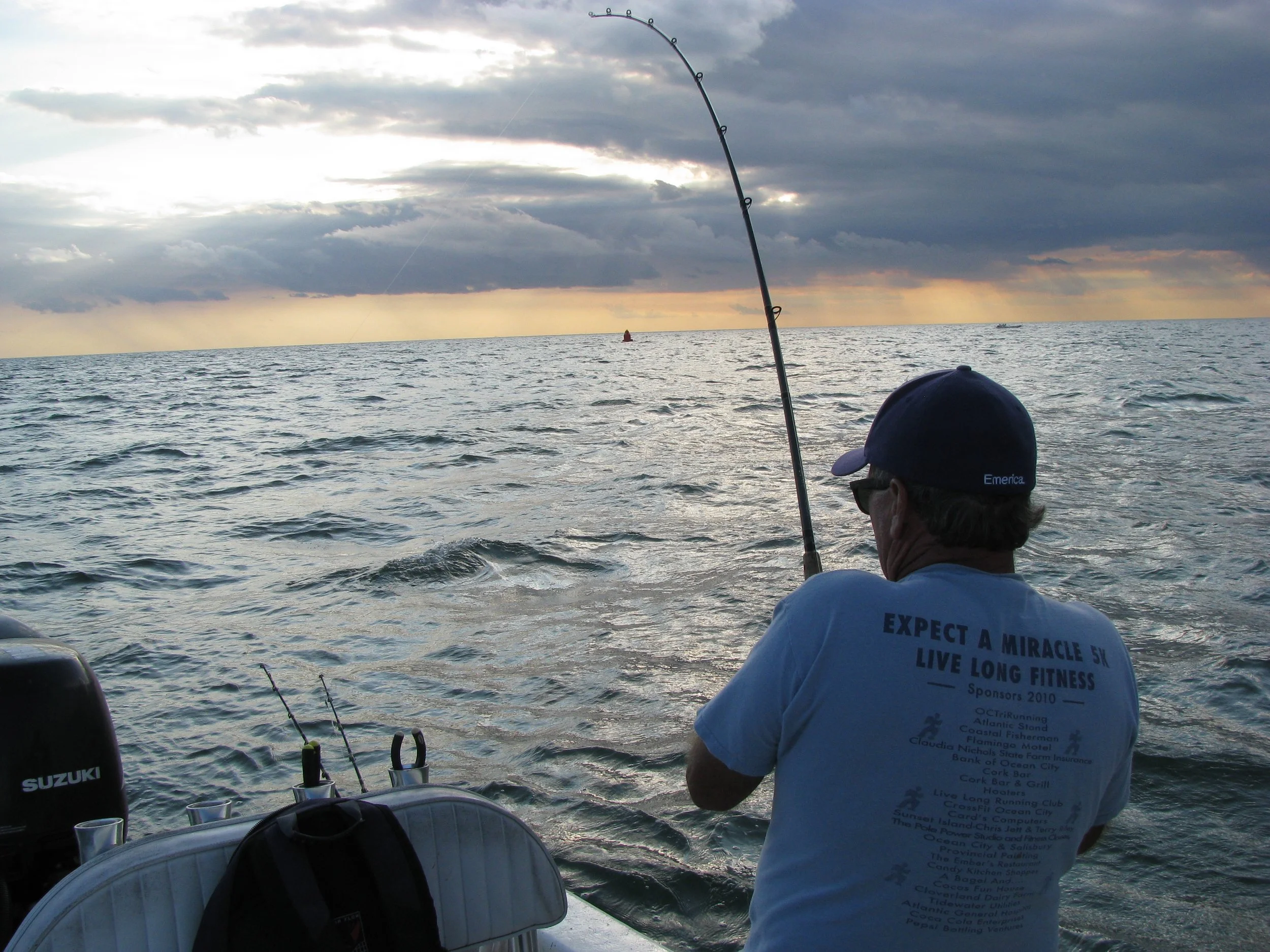 A person wearing a blue cap and white t-shirt enjoying fishing on a boat in the ocean during sunset. Fish rods are visible in the boat, and the sky is cloudy . sandbar Captain Tilly OC Fishing Charters. Ocean City, MD