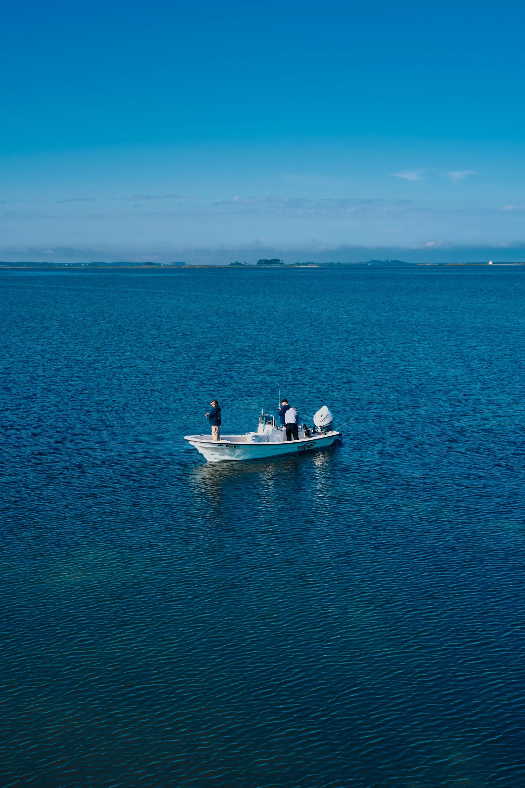 A boat with three people on it floating on a calm body of water, with a distant shoreline and a clear blue sky.