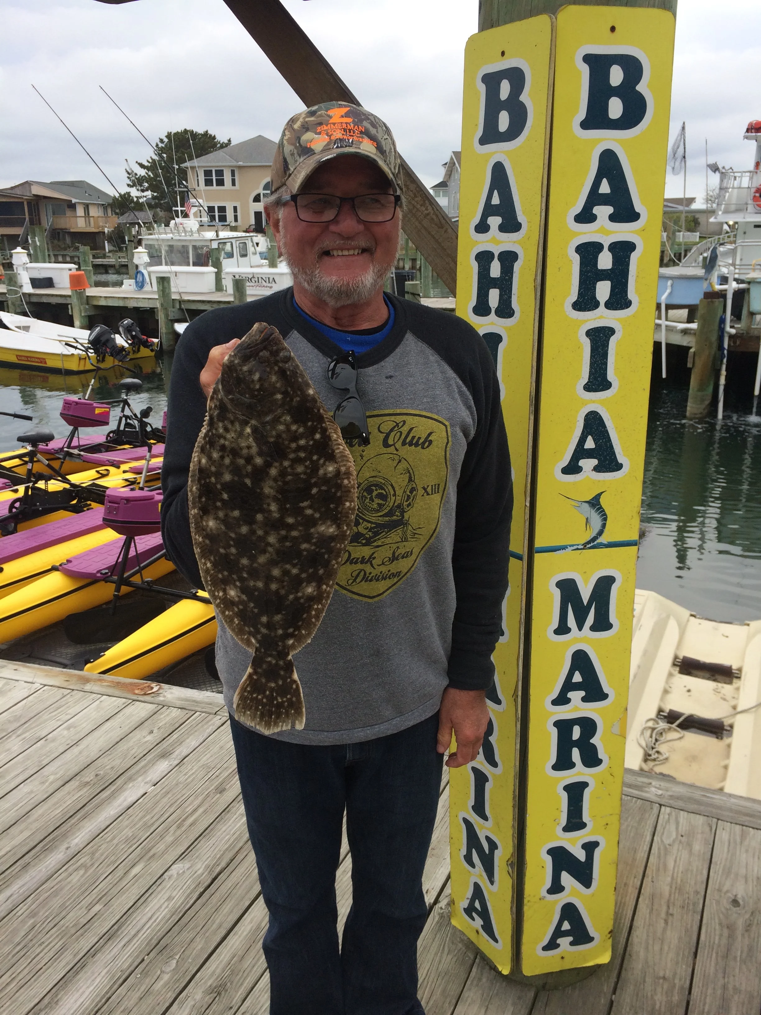 A man with glasses and a gray beard smiling while holding a flatfish in front of a yellow sign with nautical words, on a wooden dock with boats in the background. sandbar Captain Tilly OC Fishing Charters. Ocean City, MD