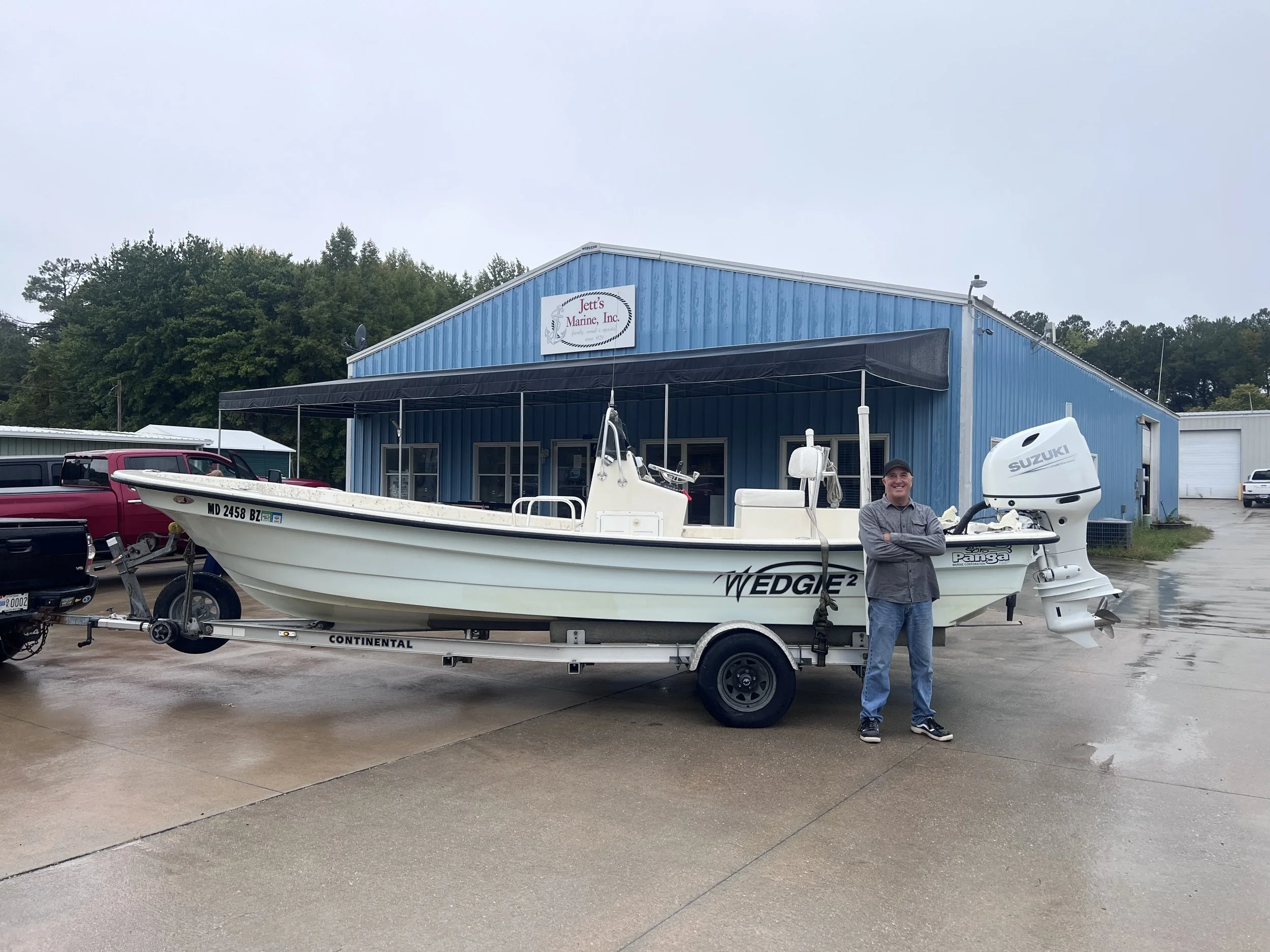 A man standing next to a white boat with a Suzuki outboard motor on a trailer in front of a blue building with a sign that reads Jett's Marine, Inc. sandbar Captain Tilly OC Fishing Charters. Ocean City, MD