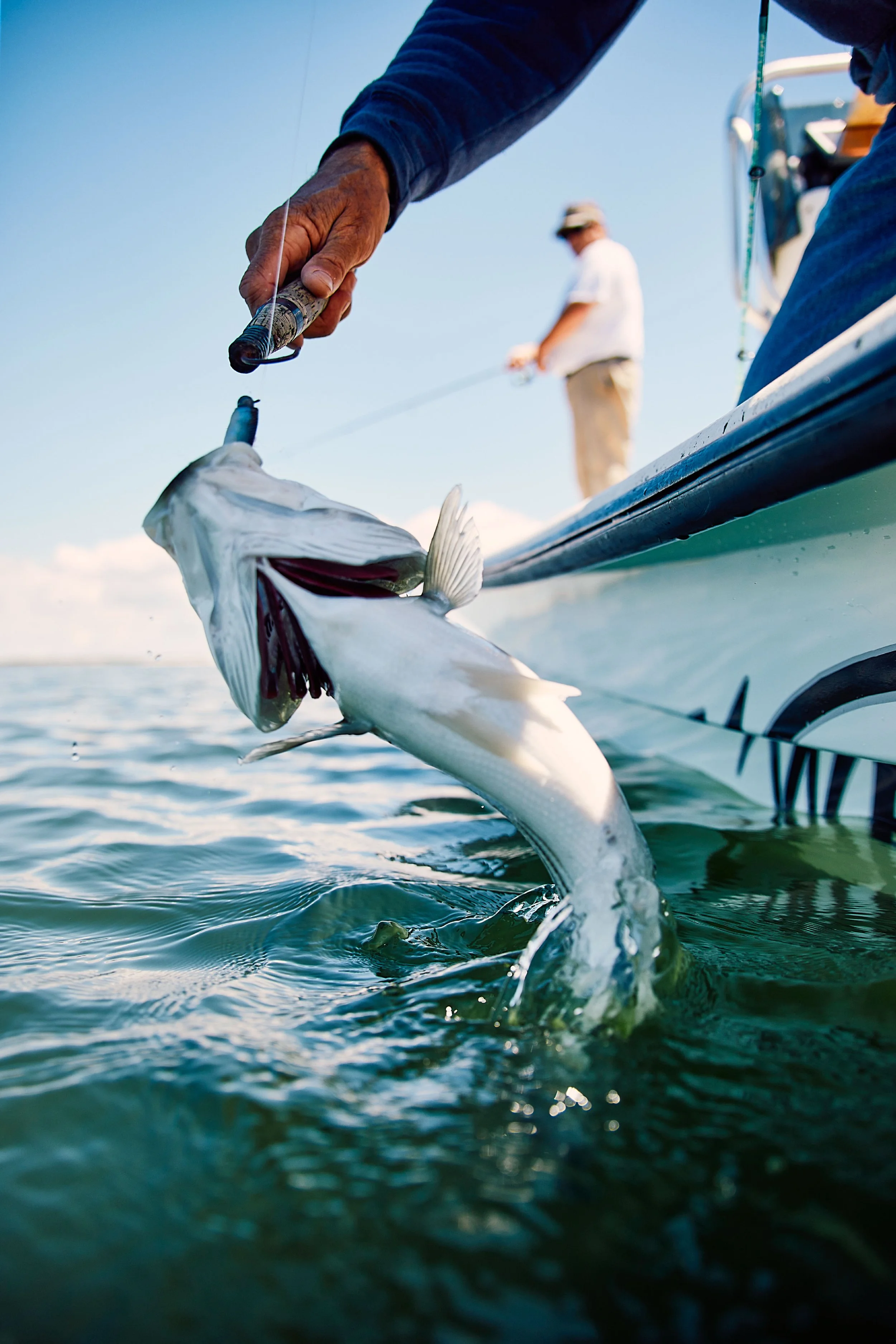 Close-up of a person reeling in a fish on a boat, with another person fishing in the background under clear blue skies. sandbar Captain Tilly OC Fishing Charters. Ocean City, MD