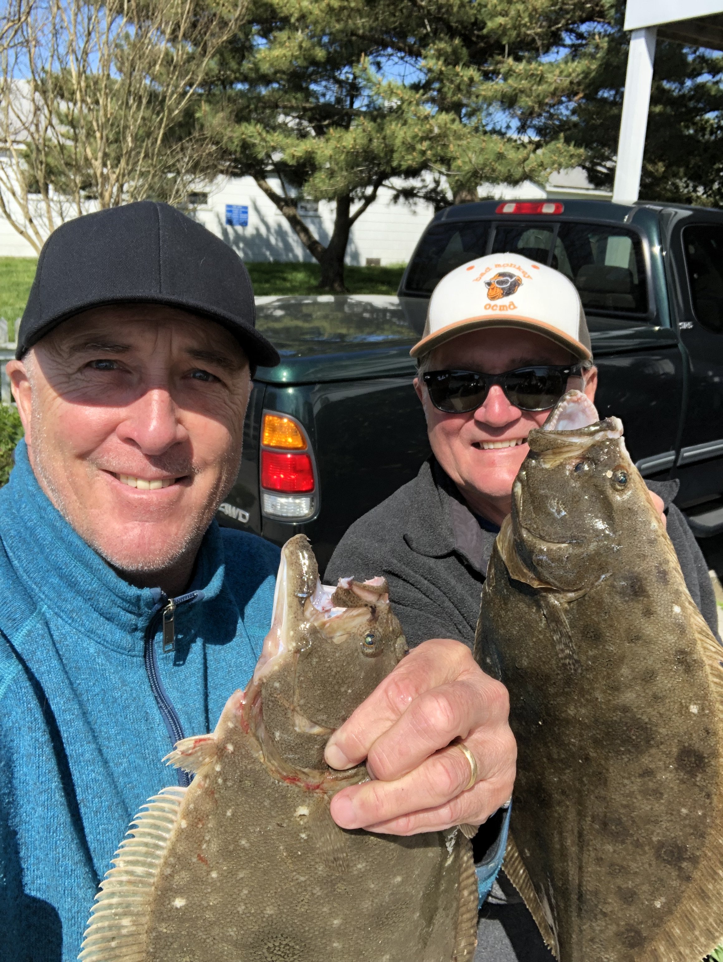 Two men smiling and holding fish, outdoors with trees and a building in the background. sandbar Captain Tilly OC Fishing Charters. Ocean City, MD