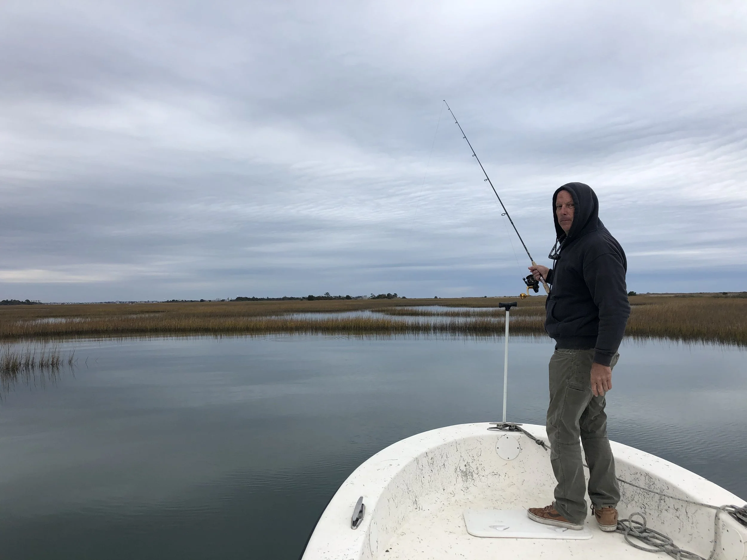 A man wearing a hoodie and cargo pants standing on the edge of a small white boat, fishing in a calm body of water with marshy land and overcast sky in the background. sandbar Captain Tilly OC Fishing Charters. Ocean City, MD