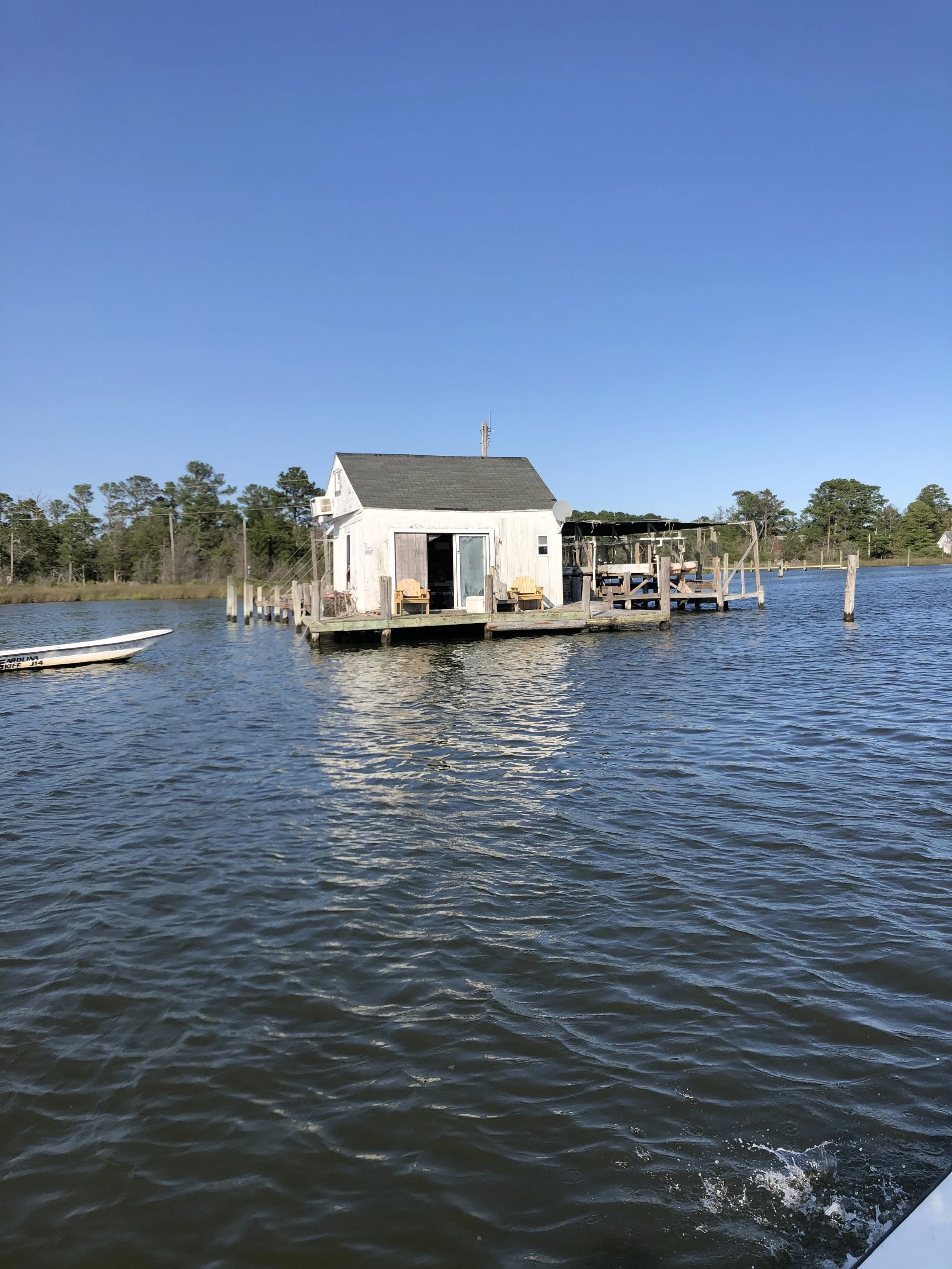 A small white wooden house on stilts over a body of water, with trees in the background and a clear blue sky above. sandbar Captain Tilly OC Fishing Charters. Ocean City, MD