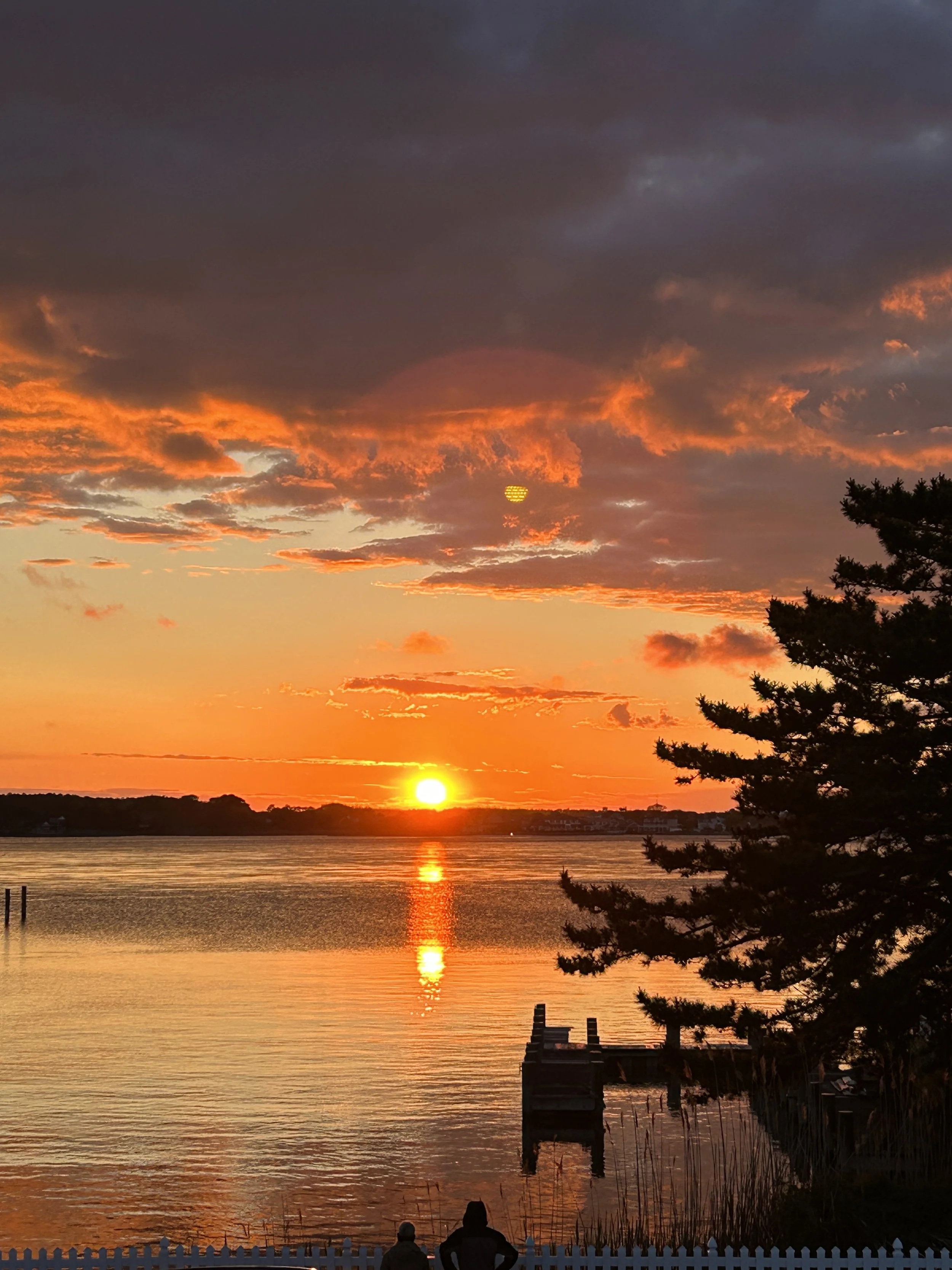 A sunset over a body of water with clouds in the sky, a silhouette of trees on the right, and two people sitting near a white picket fence at the bottom. sandbar Captain Tilly OC Fishing Charters. Ocean City, MD