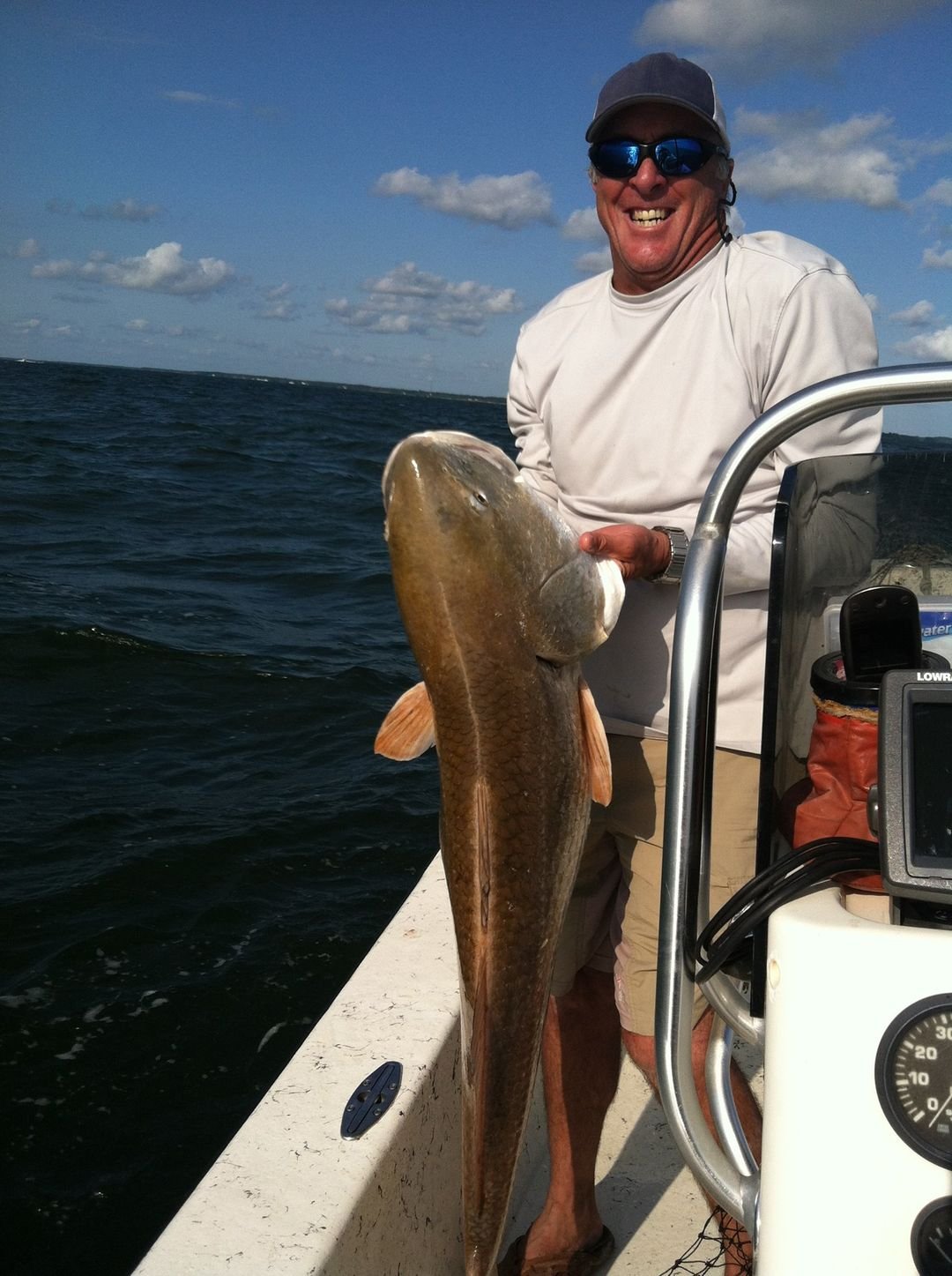 Smiling man wearing sunglasses and a baseball cap holding a large fish on a boat in the ocean with blue sky and clouds in the background. sandbar Captain Tilly OC Fishing Charters. Ocean City, MD