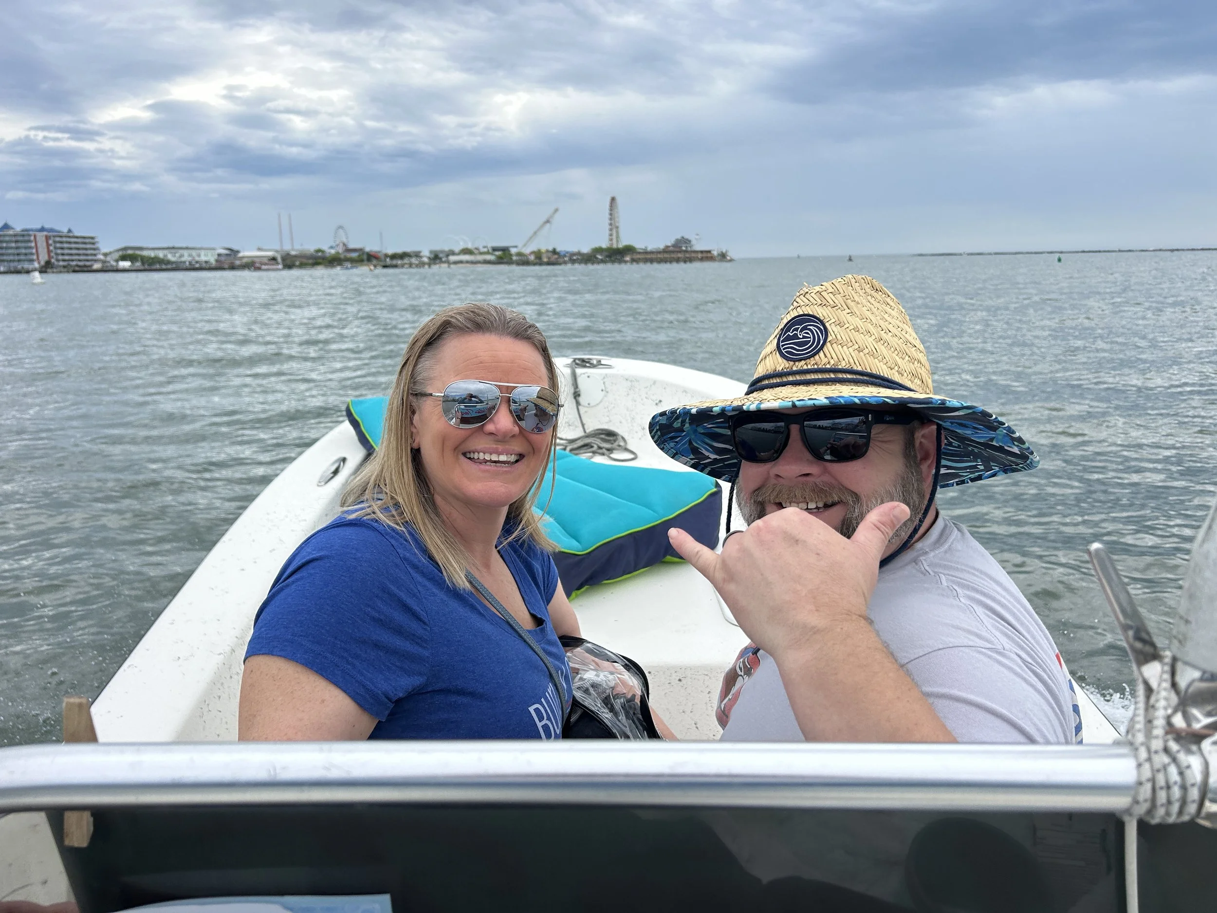 Two people on a boat taking a selfie with a water amusement park and city skyline in the background, overcast sky.