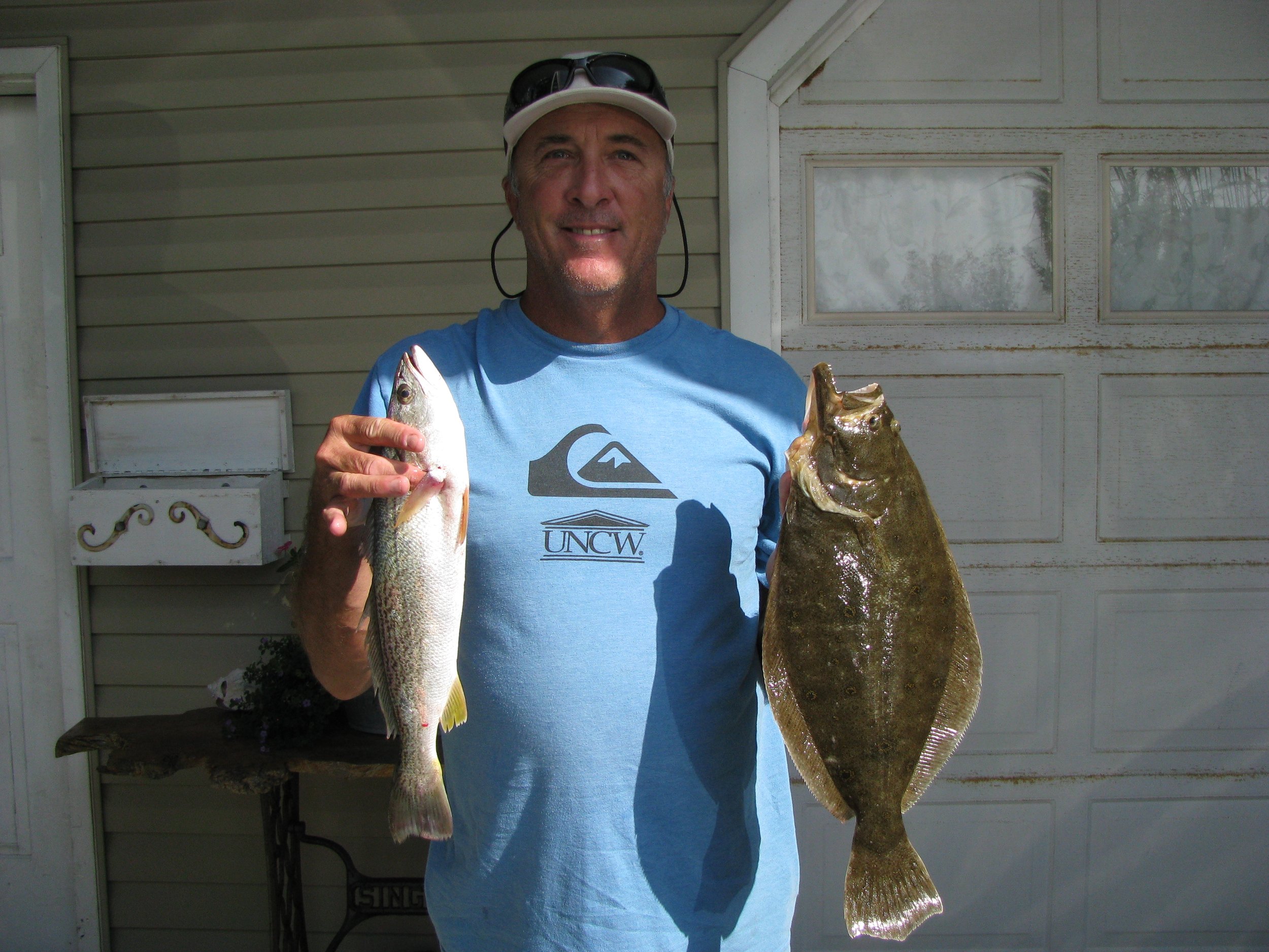 A man wearing a white cap and sunglasses, holding two fish, standing in front of a house with beige siding and a garage door. sandbar Captain Tilly OC Fishing Charters. Ocean City, MD