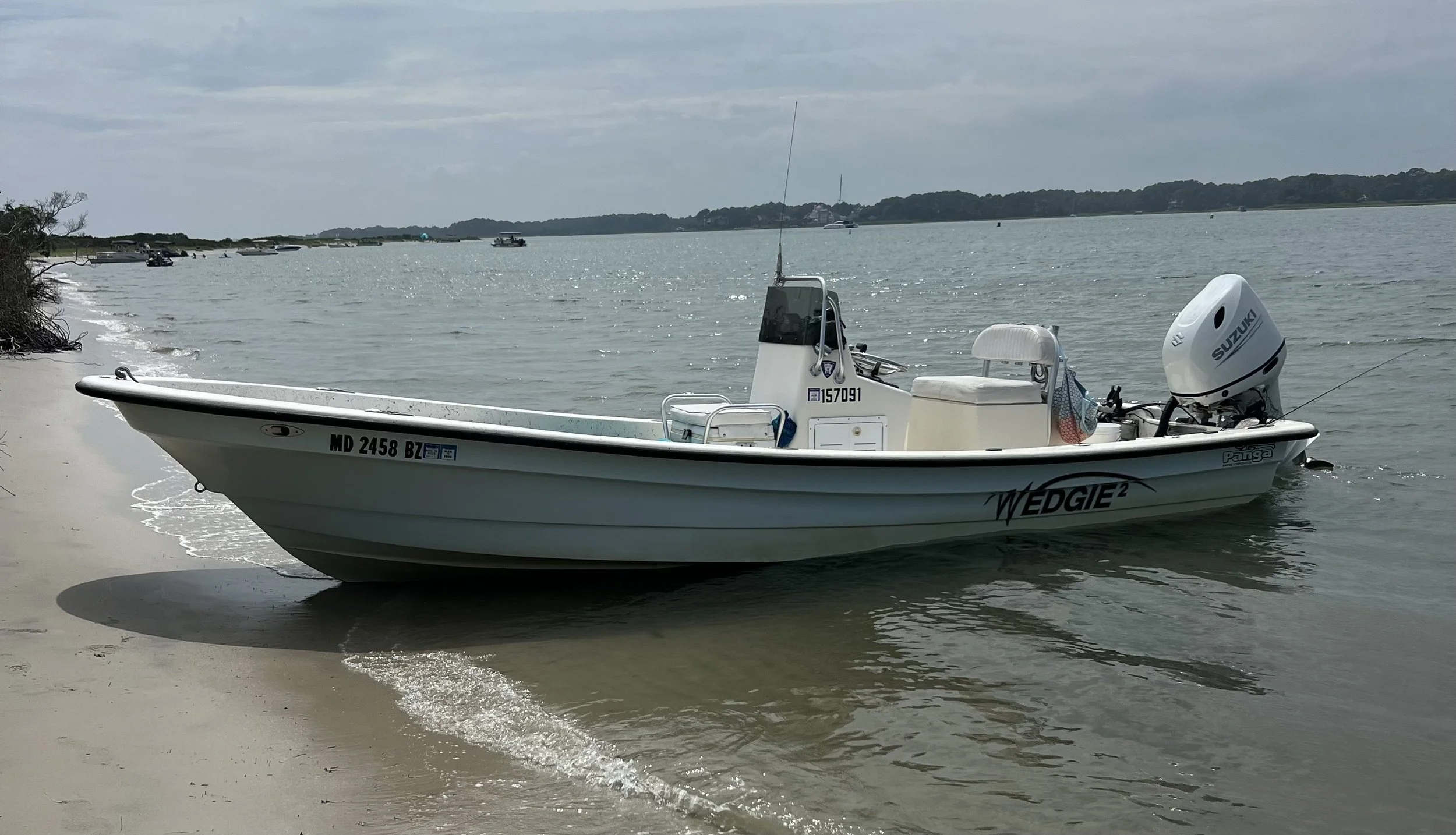 A small white boat with outboard motor and a Suzuki engine resting on a sandy shoreline, with calm water and distant boats and land in the background. sandbar Captain Tilly OC Fishing Charters. Ocean City, MD