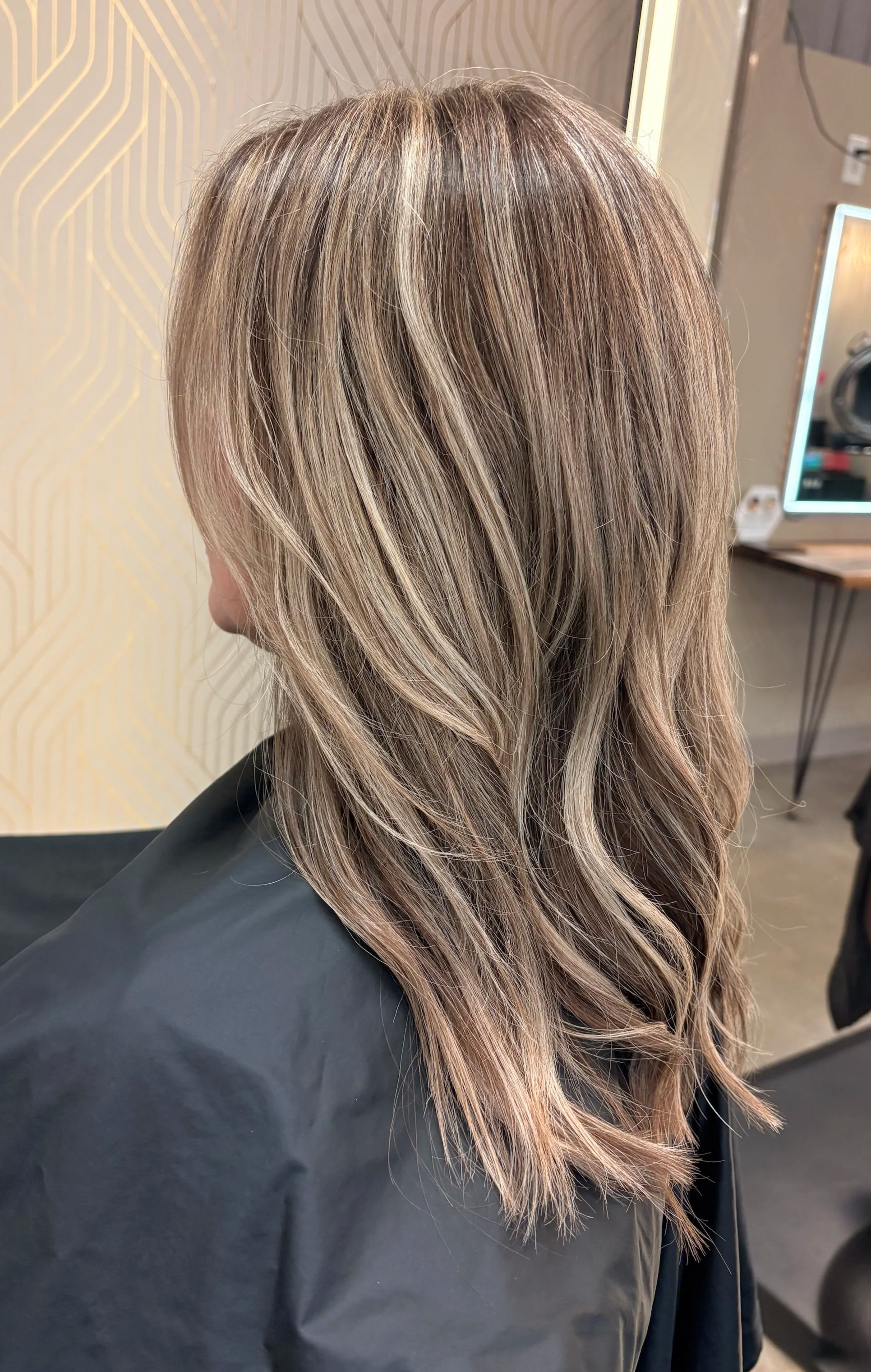 Woman with long, wavy, blonde hair sitting in a salon chair, wearing a black cape, with a decorative gold and beige patterned wall and a mirror in the background.