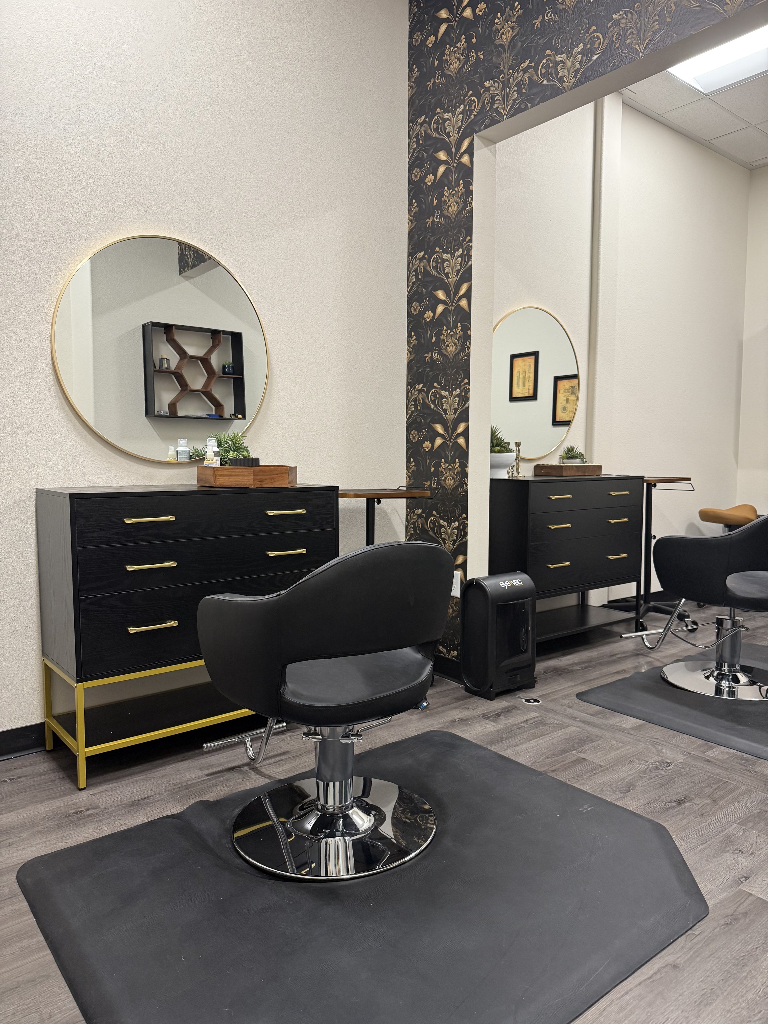 A modern hair salon in Vancouver, WA with black styling chairs, black mats, black and gold skincare cabinets, mirrors, and wallpaper with gold floral design.