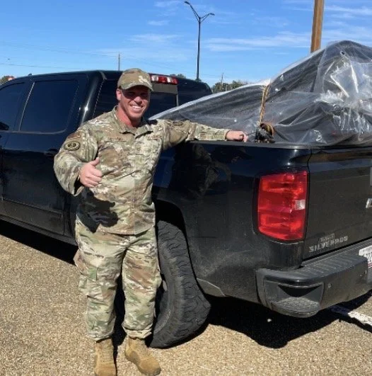 A man in military uniform giving a thumbs-up next to a black pickup truck with a large, covered object in its bed, on a dirt lot with a wind turbine in the background.