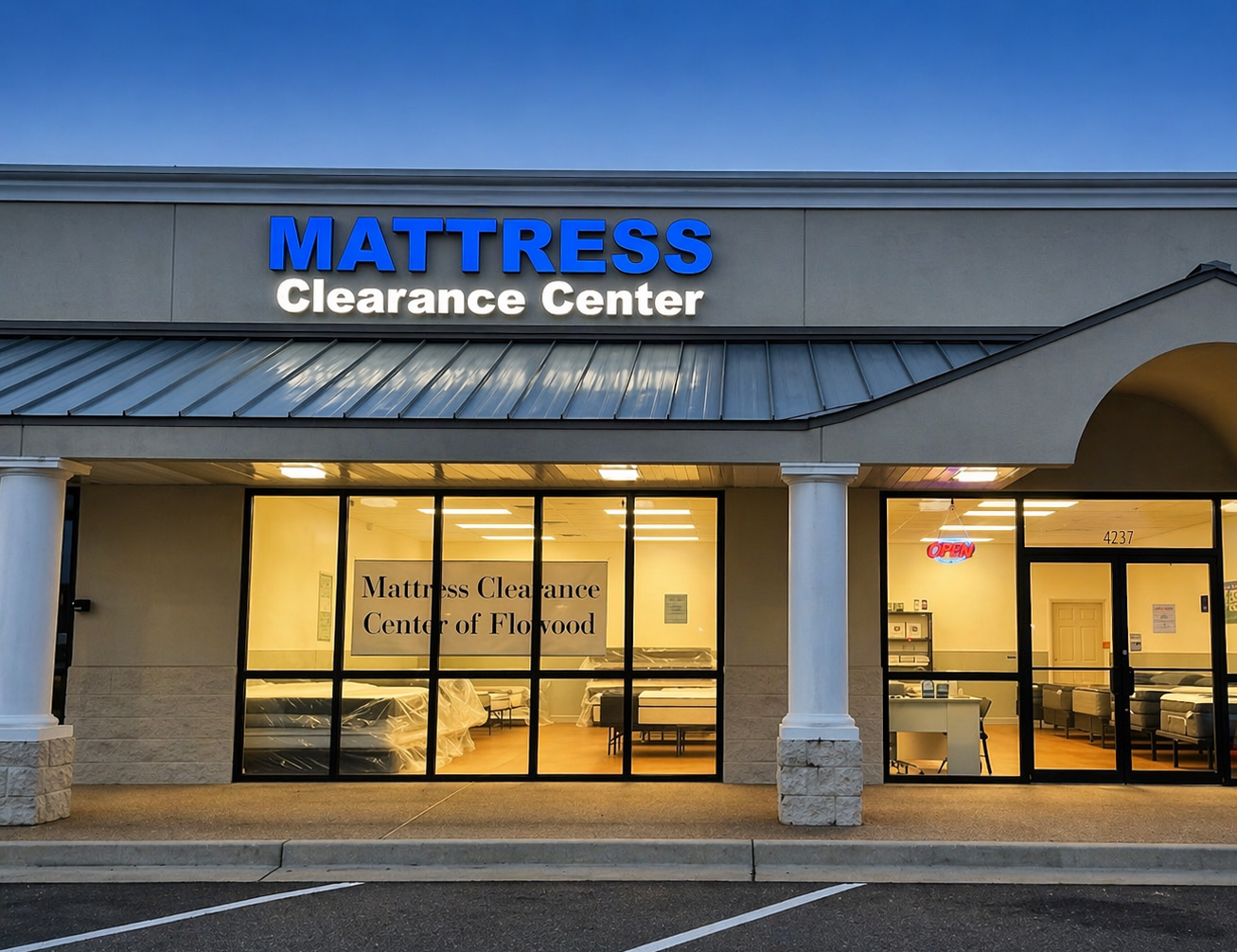 Front of a mattress store named Mattress Clearance Center, with large glass windows showing mattresses inside and an illuminated open sign. The building has a gray facade and a metal awning.
