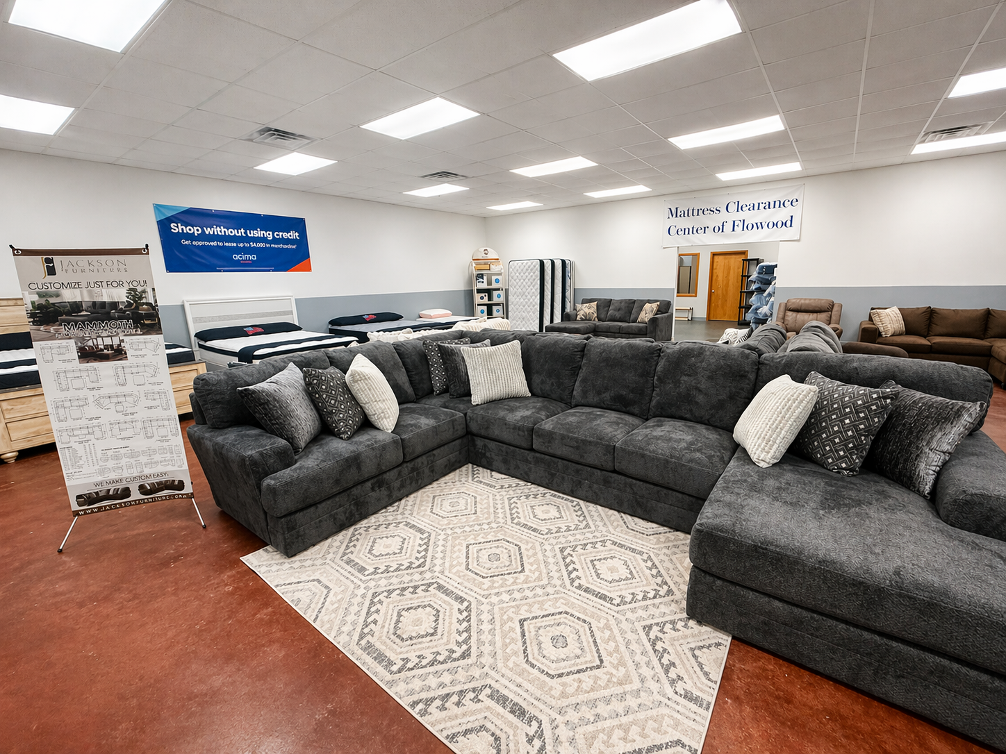 Furniture store with a large dark gray sectional sofa with multiple throw pillows on a patterned rug. In the background, there are beds, a mattress display, and signs for mattress clearance and shop financing.