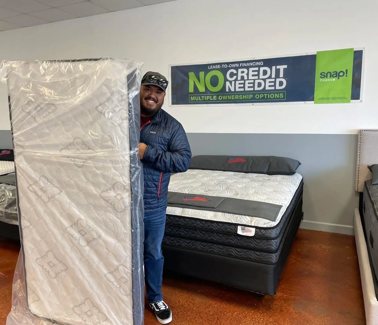 A man in a blue jacket holding a new mattress in a furniture showroom.