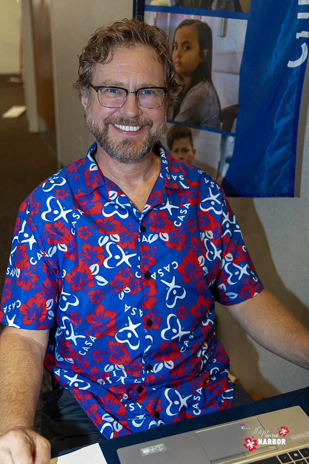 A man with glasses and a beard smiling, wearing a blue Hawaiian shirt with red and white floral and palm tree patterns, sitting at a table with a laptop in front of him, with a poster of a woman and a child in the background.