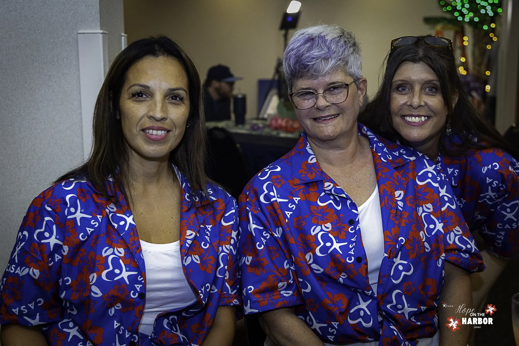 Three women wearing matching blue shirts with red and white patterns smiling at an event.