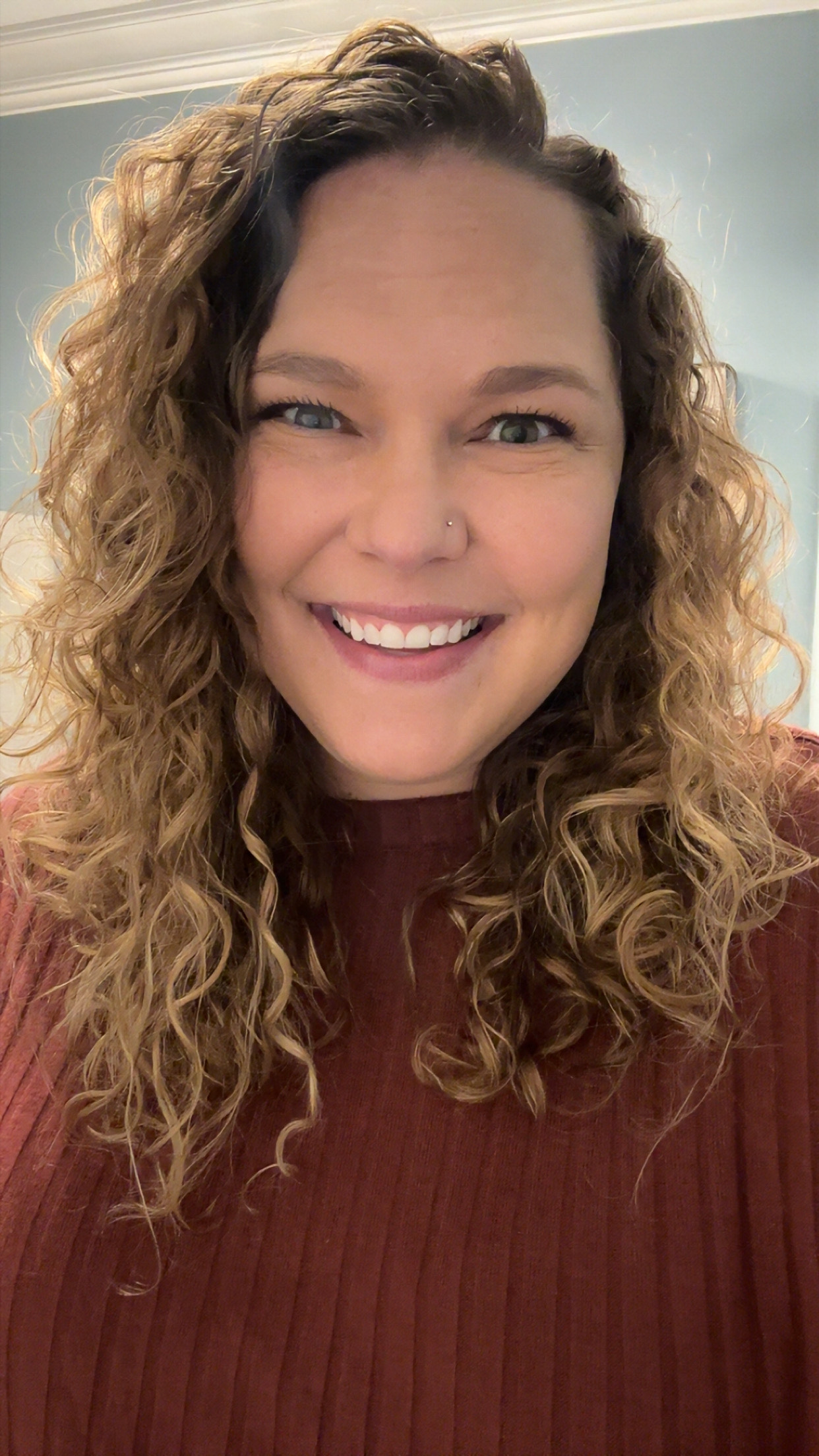 A woman with curly brown hair smiling and winking at the camera, wearing a maroon sweater.