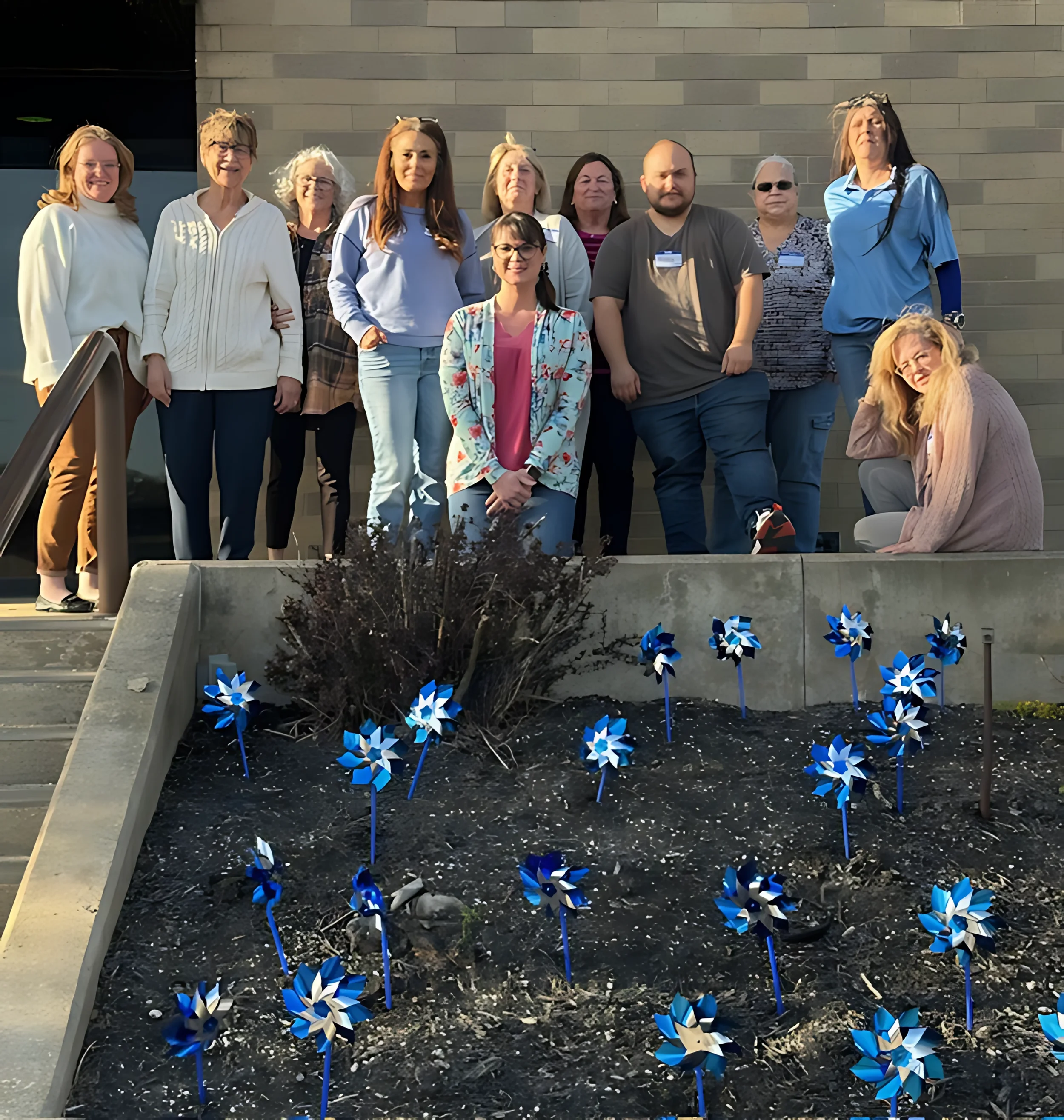 A group of people posing outdoors on steps, with some standing and one woman sitting in front, in front of a gray wall. In the foreground, there is a flower bed with blue and white pinwheel decorations.