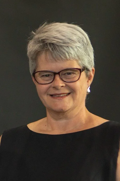A woman with short gray hair, wearing glasses and a black sleeveless top, smiling against a dark background.