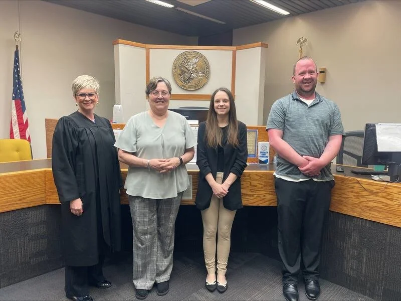 Four individuals standing in a courtroom in front of a judge's bench. The person on the left is a woman in a black judge's robe. Next to her are two women and a man, all dressed in casual or business casual attire.