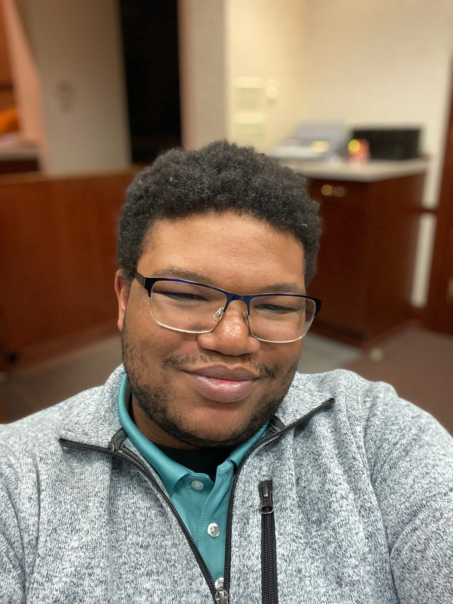 A smiling man with dark hair, glasses, and a beard, wearing a gray zip-up jacket and a teal shirt, taking a selfie in a room with wooden furniture and a blurred background.