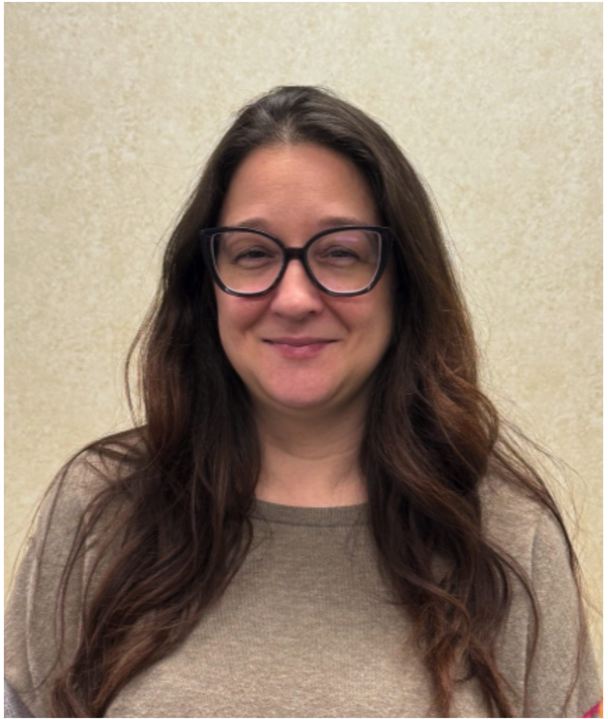 A woman with long wavy brown hair, wearing glasses and a beige shirt, standing against a plain light-colored wall.