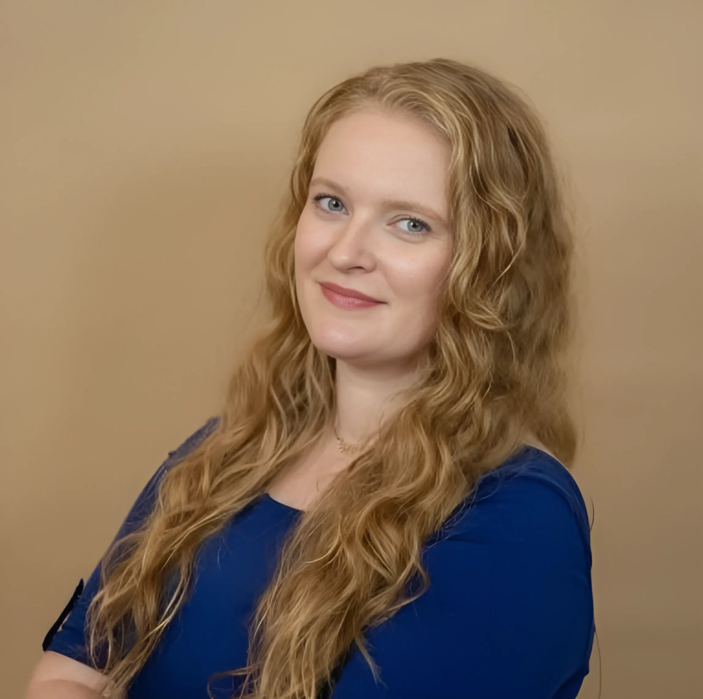 Close-up portrait of a woman with long, wavy blonde hair, wearing a blue top and smiling slightly with a beige background.