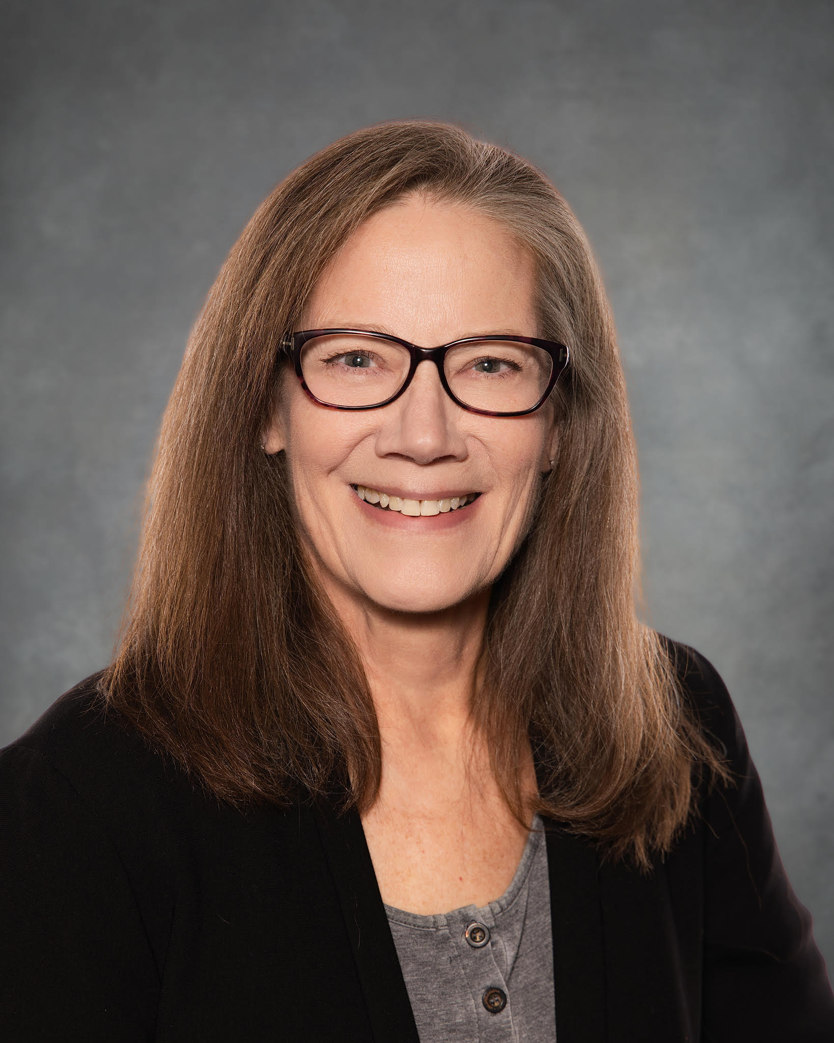 Close-up of a smiling woman with brown hair and glasses, wearing a grey top and black blazer, against a grey background.