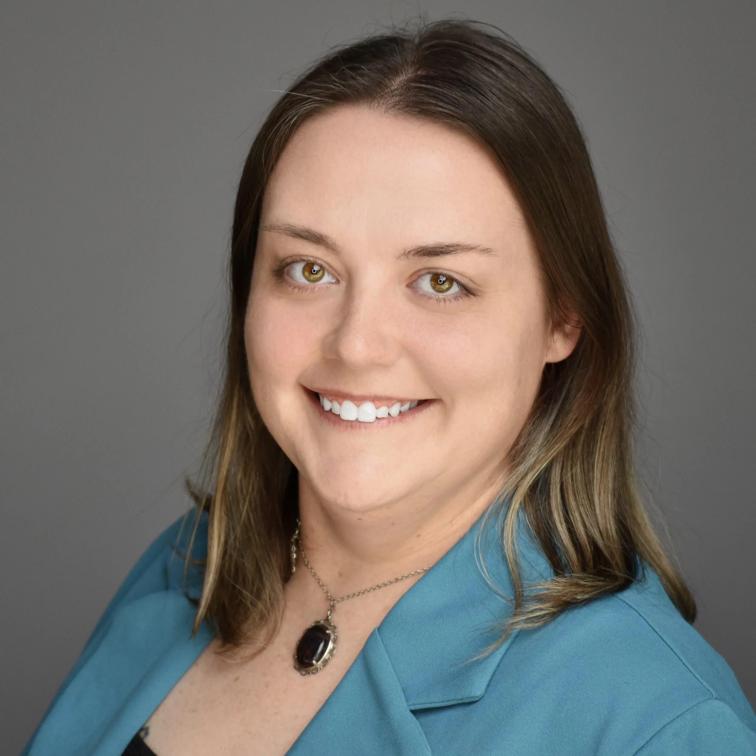 Close-up of a smiling woman with chestnut hair, green eyes, wearing a turquoise blazer and a silver necklace with a large, dark gemstone pendant, against a gray background.