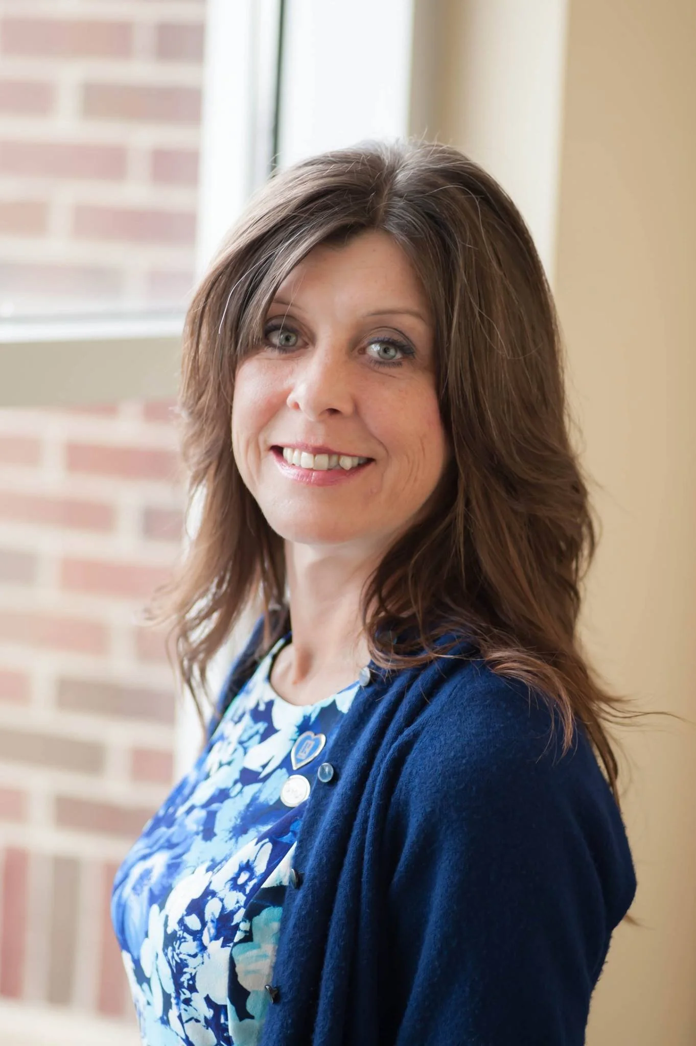 A woman with long, wavy brown hair wearing a blue patterned dress and a dark blue cardigan, smiling indoors near a window with brick wall in the background.
