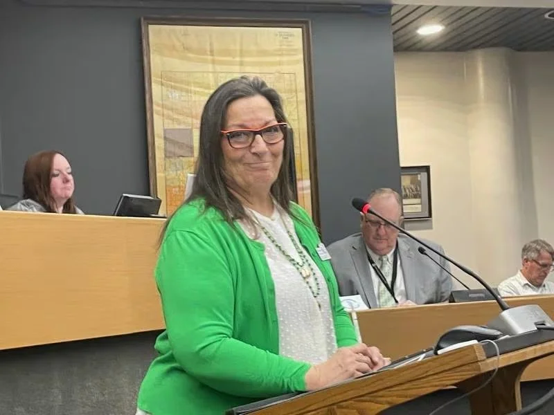 A woman with glasses smiling at a podium during a formal meeting or hearing, with a panel of people seated behind her and a large framed picture on the wall.