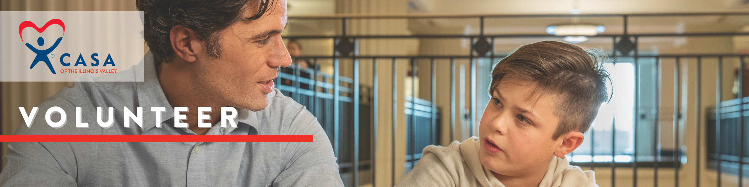 A man and a boy are having a conversation inside a building with lockers in the background. The man is wearing a light gray shirt, and the boy has a short haircut and is wearing a beige hoodie. The scene is part of a volunteer program for CASA of the Illinois Valley.