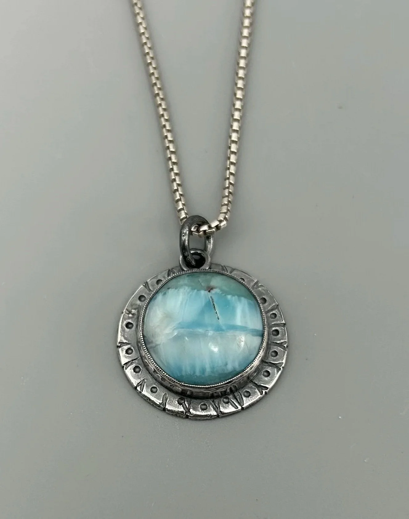 Silver necklace with a round pendant featuring a light blue gemstone. The pendant has engravings around its circumference and hangs from a silver chain.