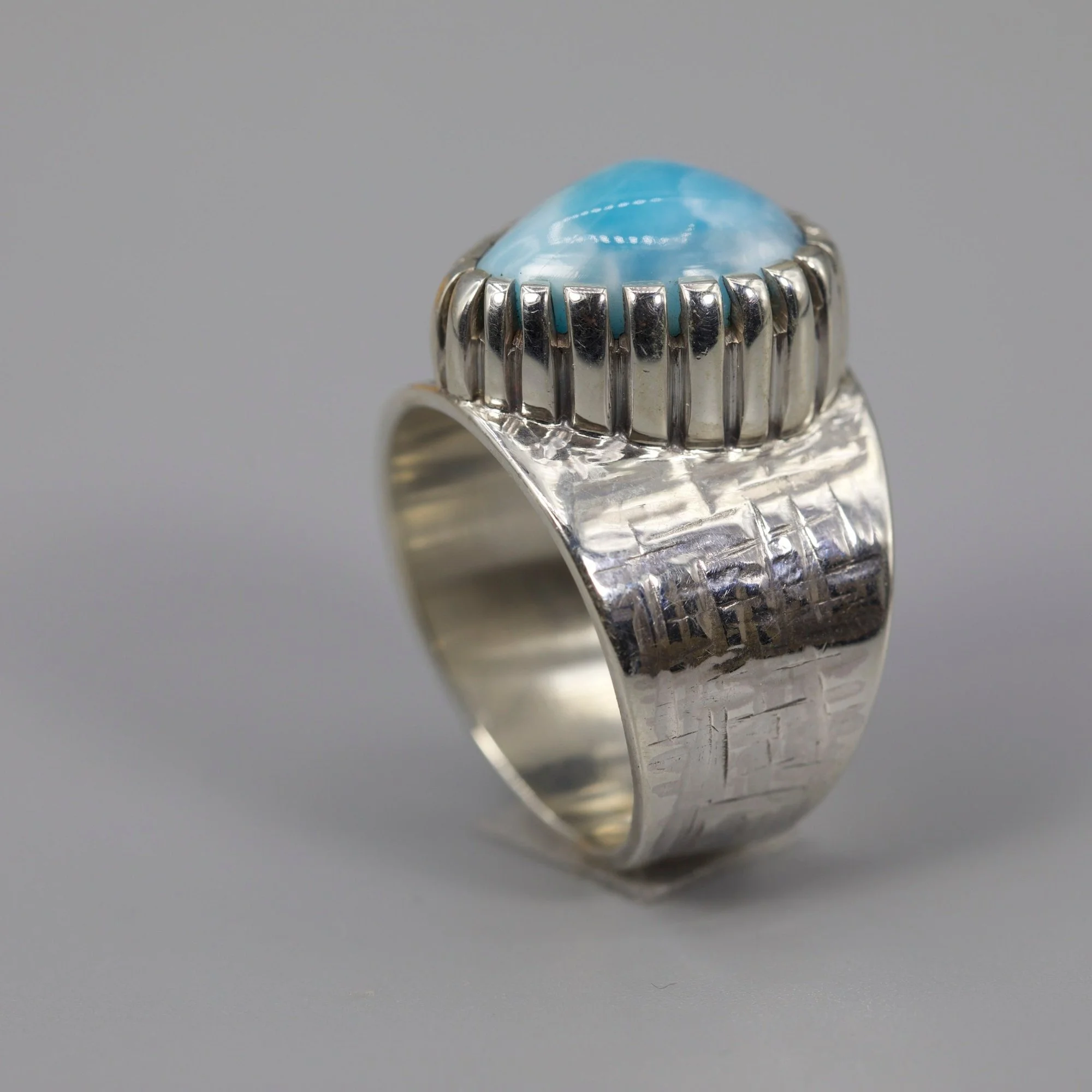 Close-up of a silver ring with a large, oval blue gemstone and vertical ridges on the band, set against a plain gray background.