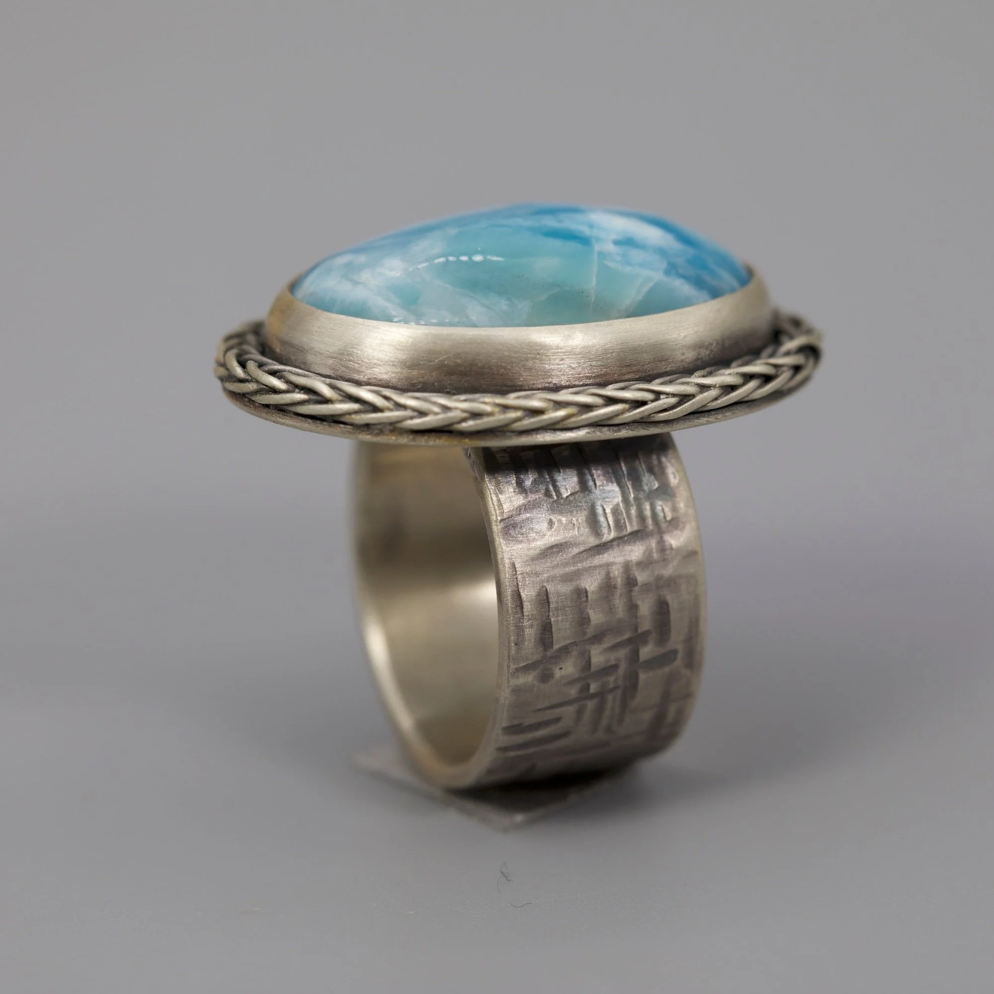 A silver ring with a large, oval-shaped blue gemstone set in the center. The gemstone has a marbled pattern with lighter and darker blue shades. The ring has a decorative braided or twisted design around the setting, and the band has a textured, patt