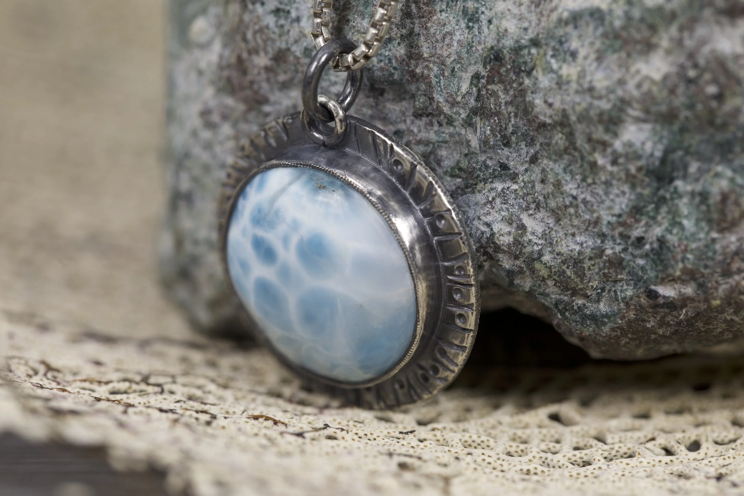 A close-up photo of a jewelry necklace with a circular charm, featuring a blue and white sky scene, hanging on a textured stone surface.