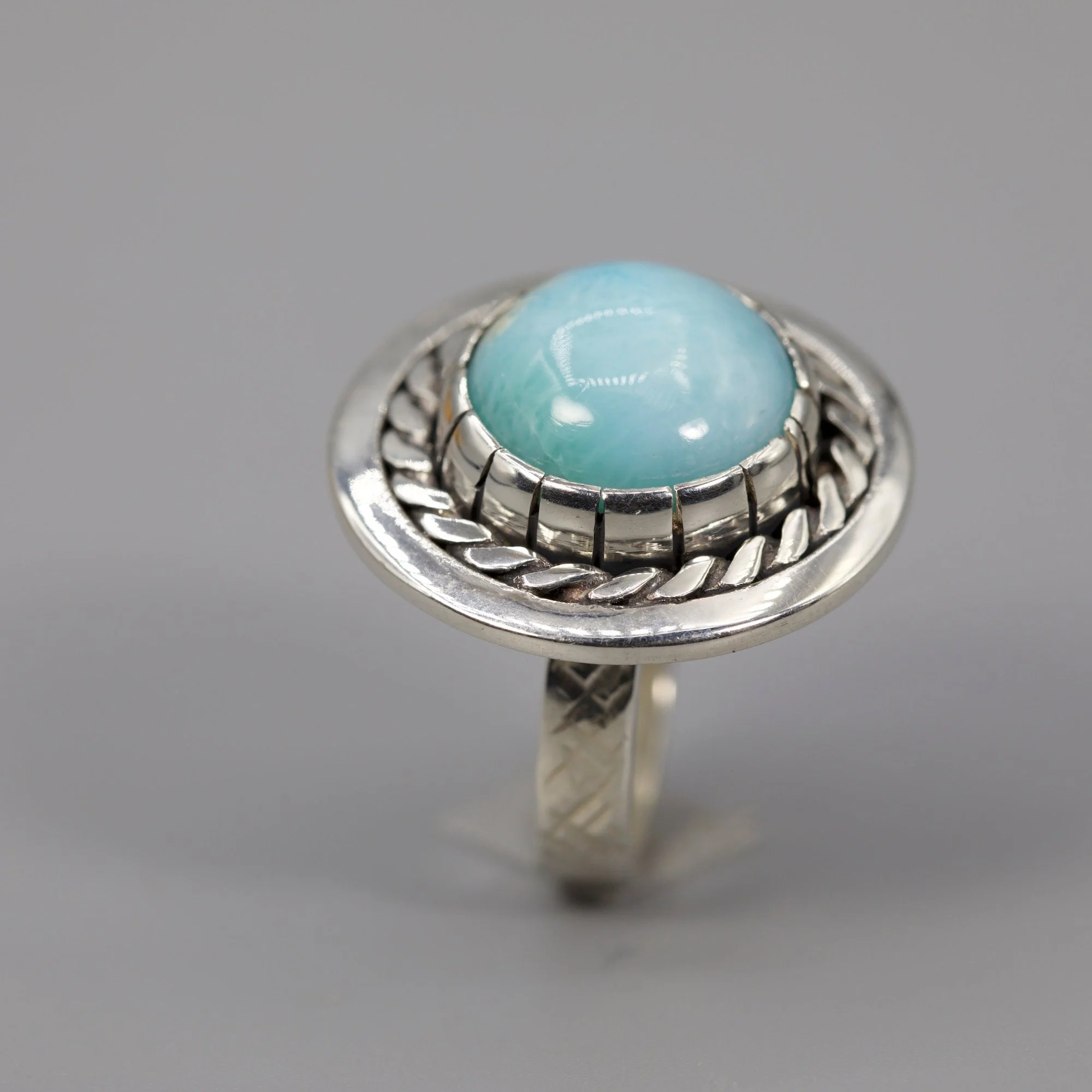 A silver ring with a light blue gemstone set in a detailed band, displayed against a plain gray background.