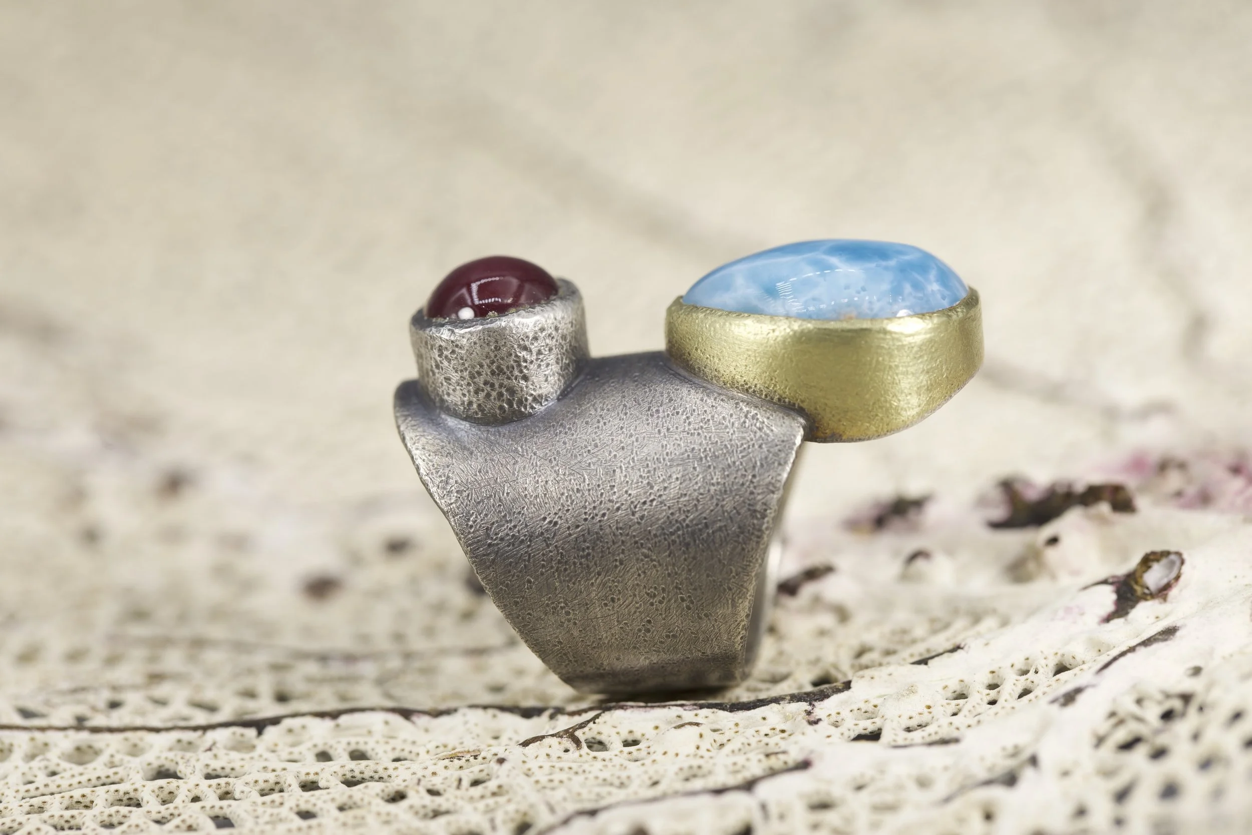 Close-up photo of a silver and gold ring with a red and a blue gemstone, resting on a textured white surface.