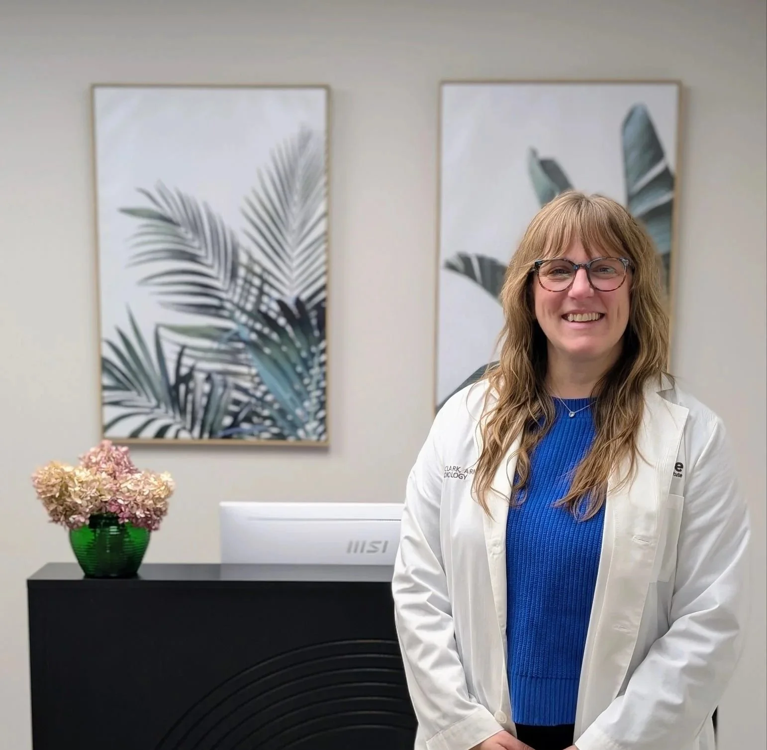 Provider NP Darcie Clark wearing glasses and a white lab coat standing in an office or clinic, smiling. Behind her, there are two framed prints on the wall, a black cabinet with a green vase of pink flowers on top, and a white computer monitor.