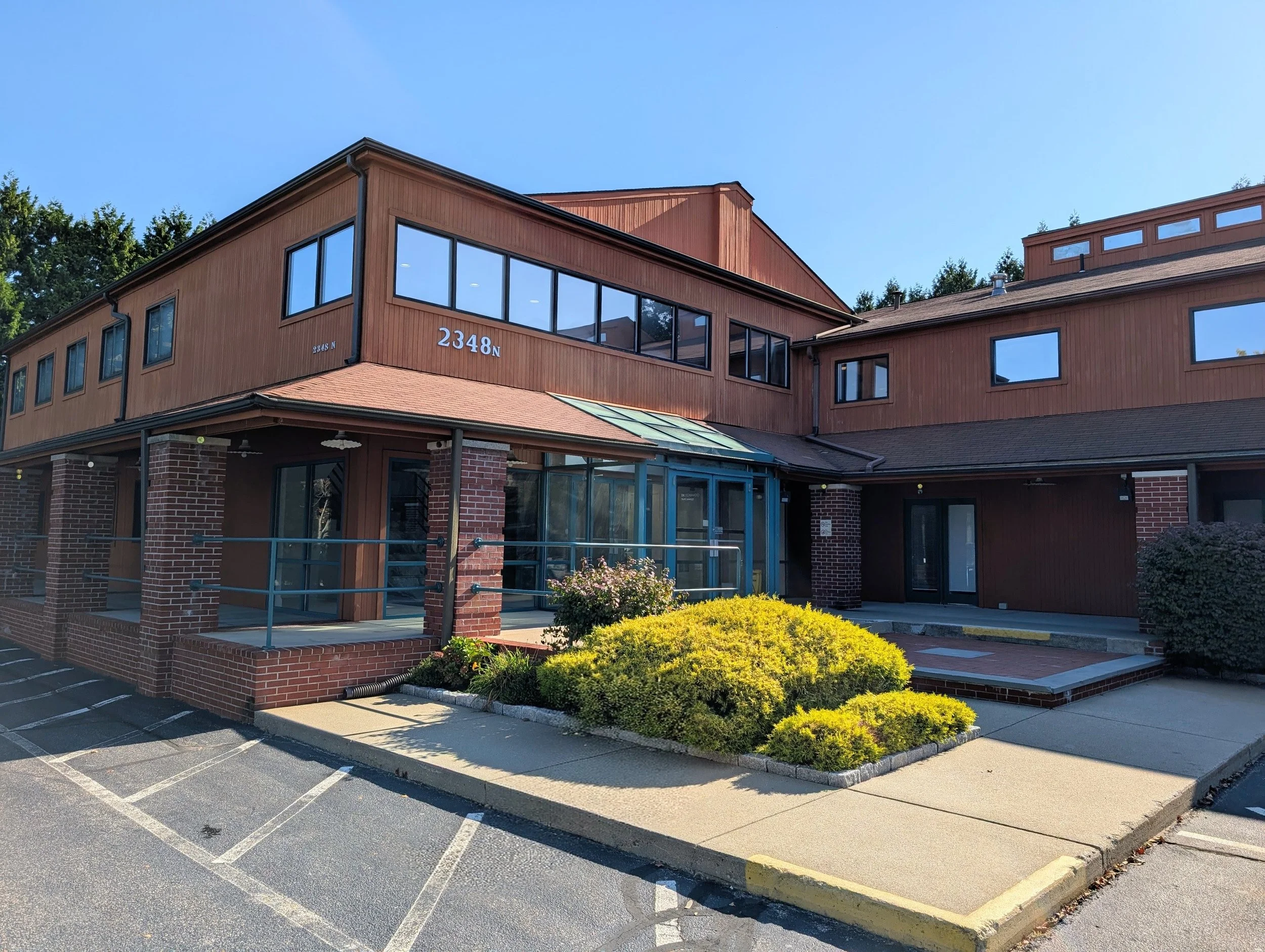 A multi-story building with a wood siding exterior, large windows, brick accents, and a parking lot with marked parking spaces in the foreground. There are green bushes and a small landscaped area at the base of the building.