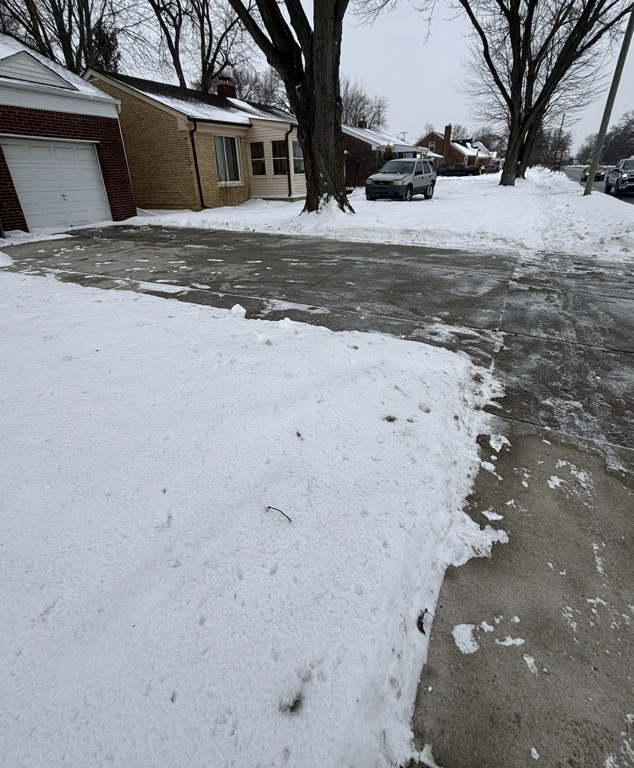 Snow-covered residential neighborhood with a brick and siding house, leafless trees, a parked silver vehicle, and a sidewalk with snow and cleared patches.