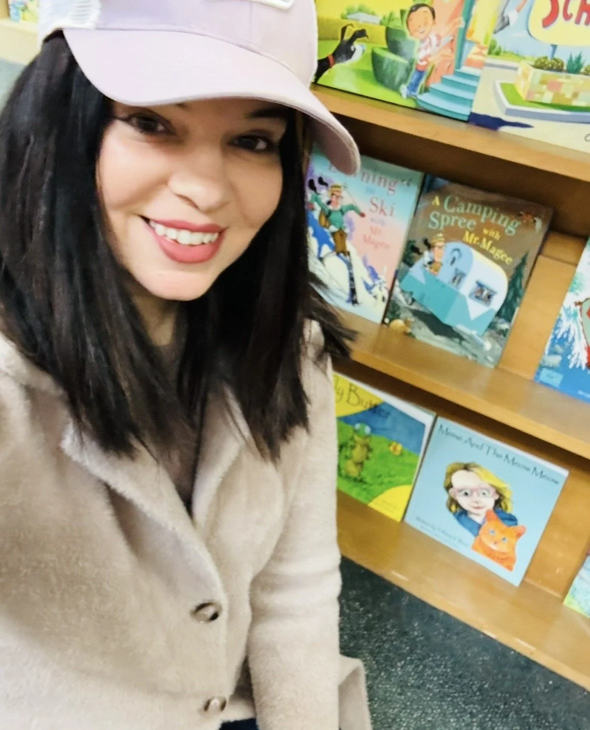 A woman with dark hair and a beige coat taking a selfie in a bookstore. Behind her are colorful children's books on wooden shelves, including titles about skiing, camping, and a girl with a cat.