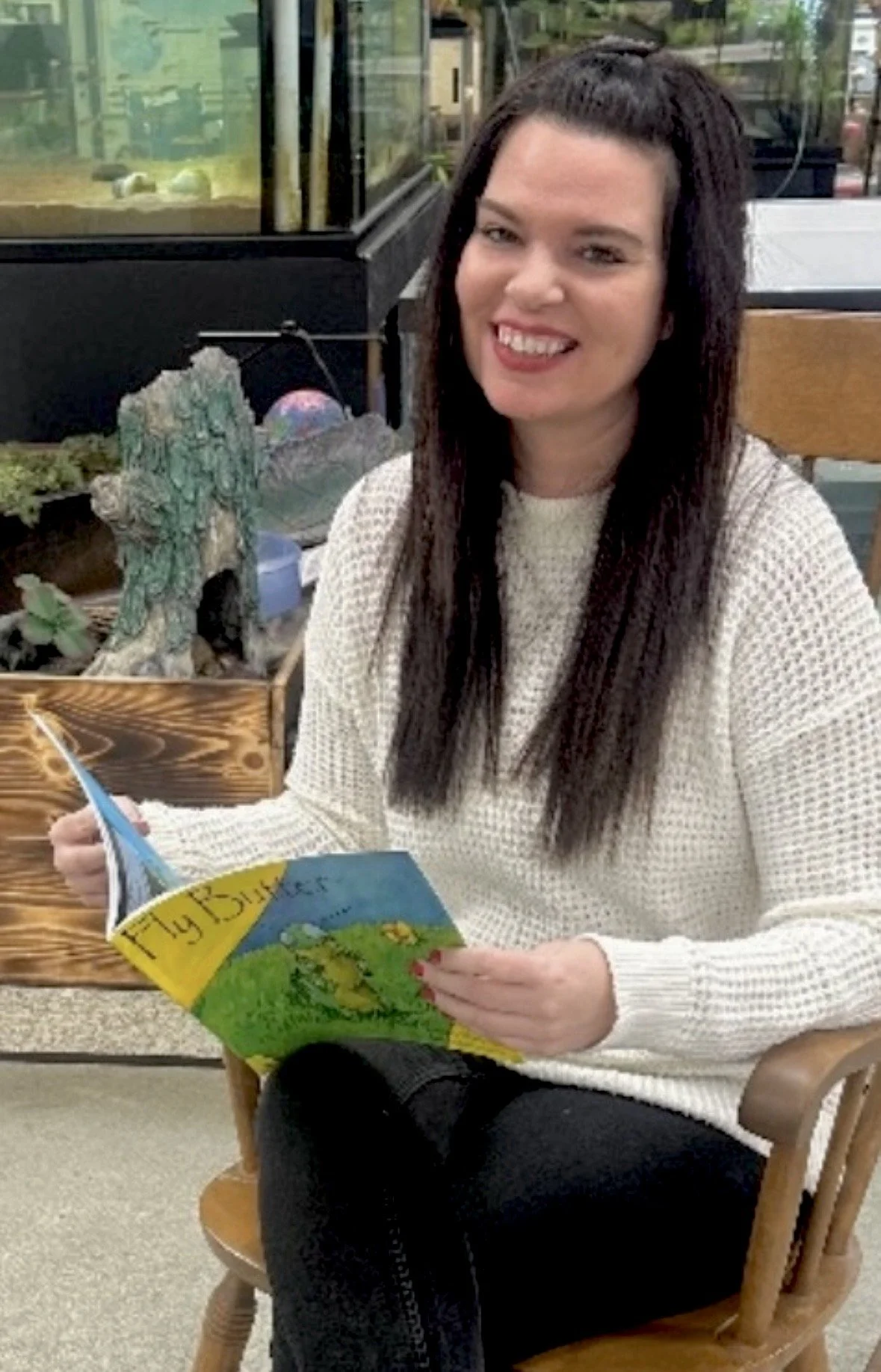 A woman with long dark hair, sitting on a wooden chair, smiling, and holding a children's book titled "Fly Butterfly" in a room with a large glass aquarium and a decorative rock arrangement in the background.