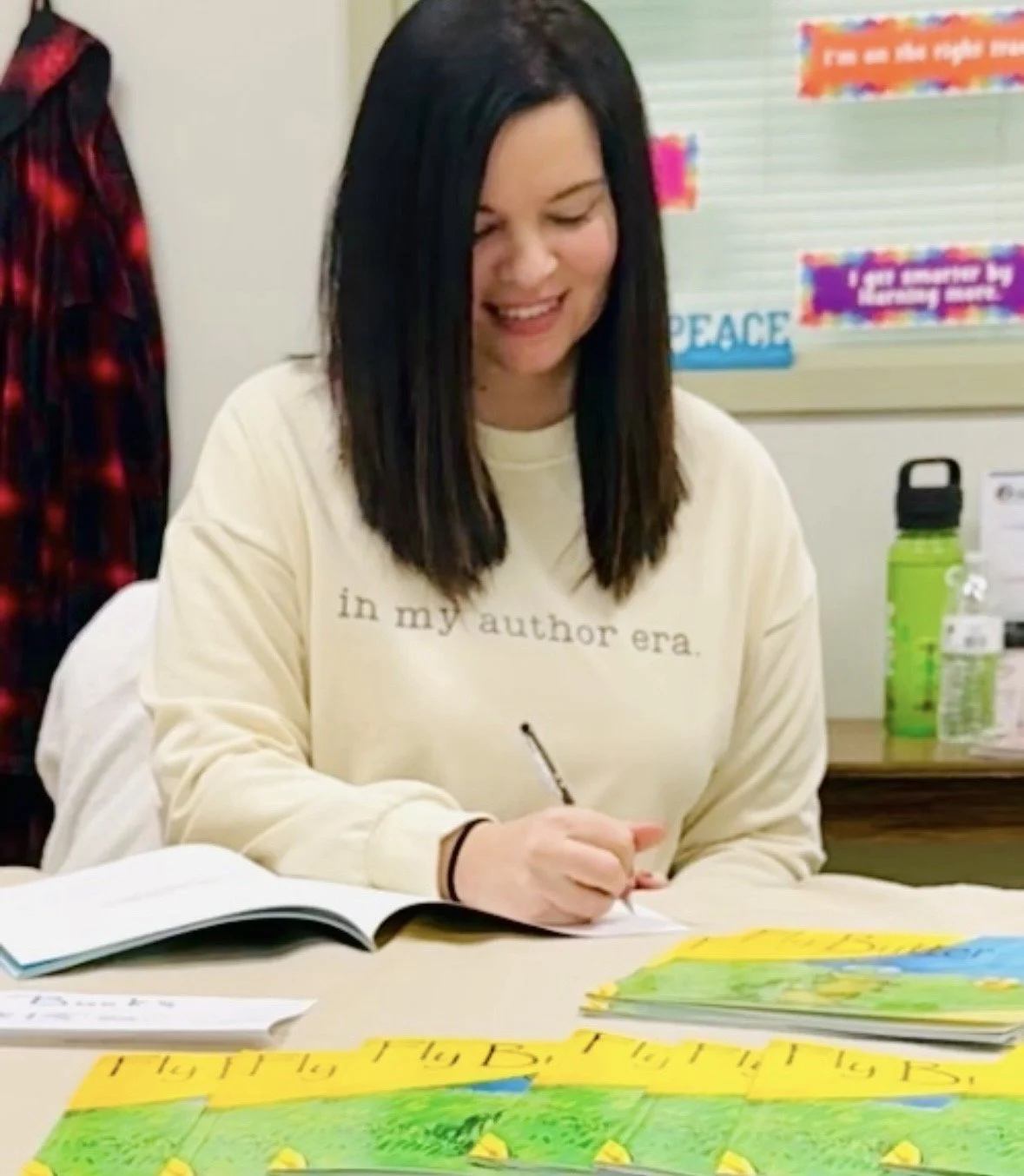 A woman with shoulder-length dark hair smiling while writing on paper at a table with colorful artwork and books. She is wearing a beige sweatshirt with text that reads 'in my author era.' Behind her, there is a bulletin board with colorful decorations and a green water bottle.