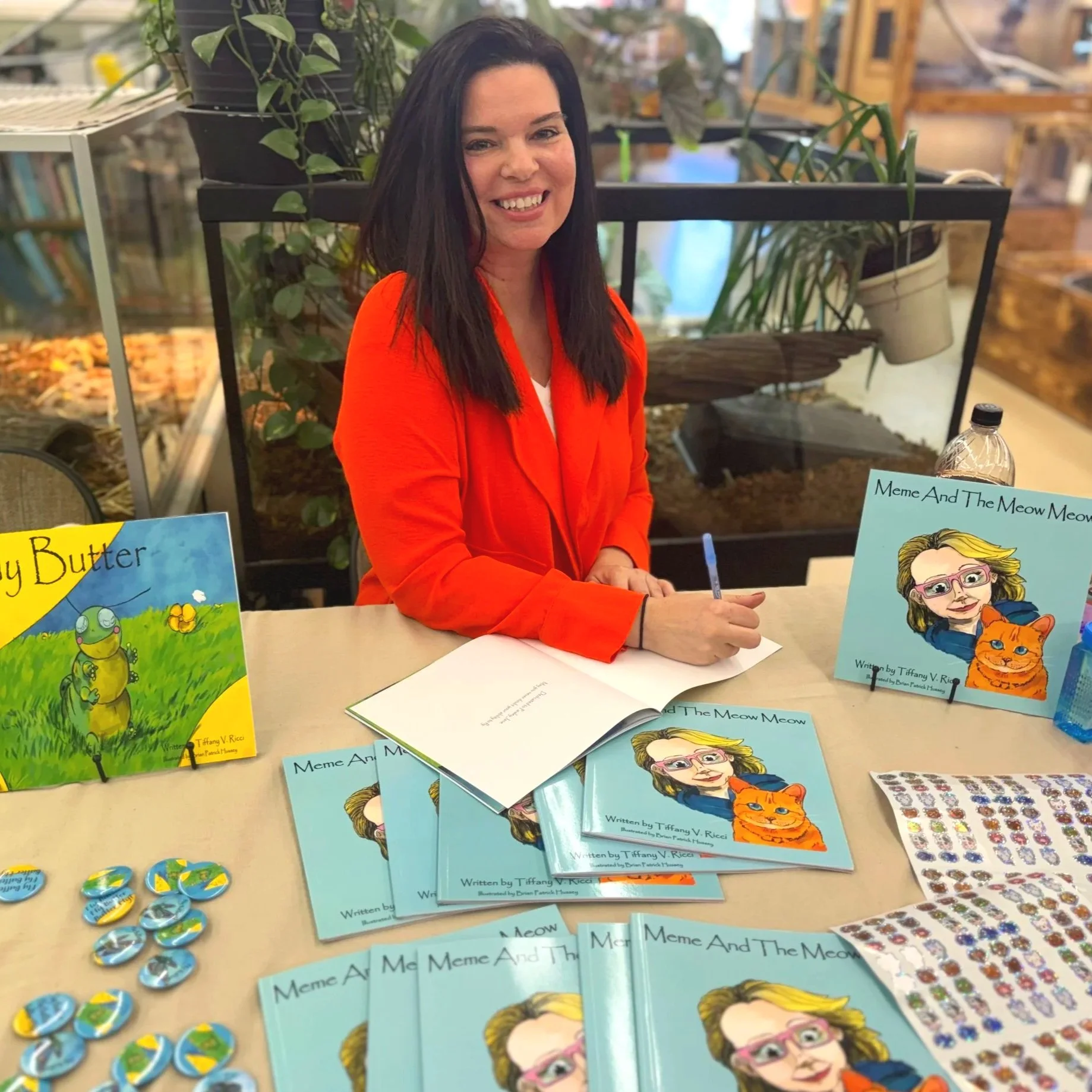 A woman with long dark hair, smiling, seated at a table displaying children's books titled "Meme And The Meow Meow" with colorful illustrations of a girl and a cat on the covers. There are also stickers and small items on the table. Behind her, there are indoor plants and bookstore shelves.