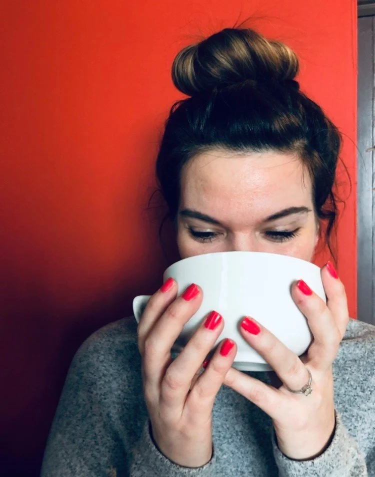 A woman with dark hair in a bun is holding a white mug close to her face, with her eyes closed, in front of a red wall.