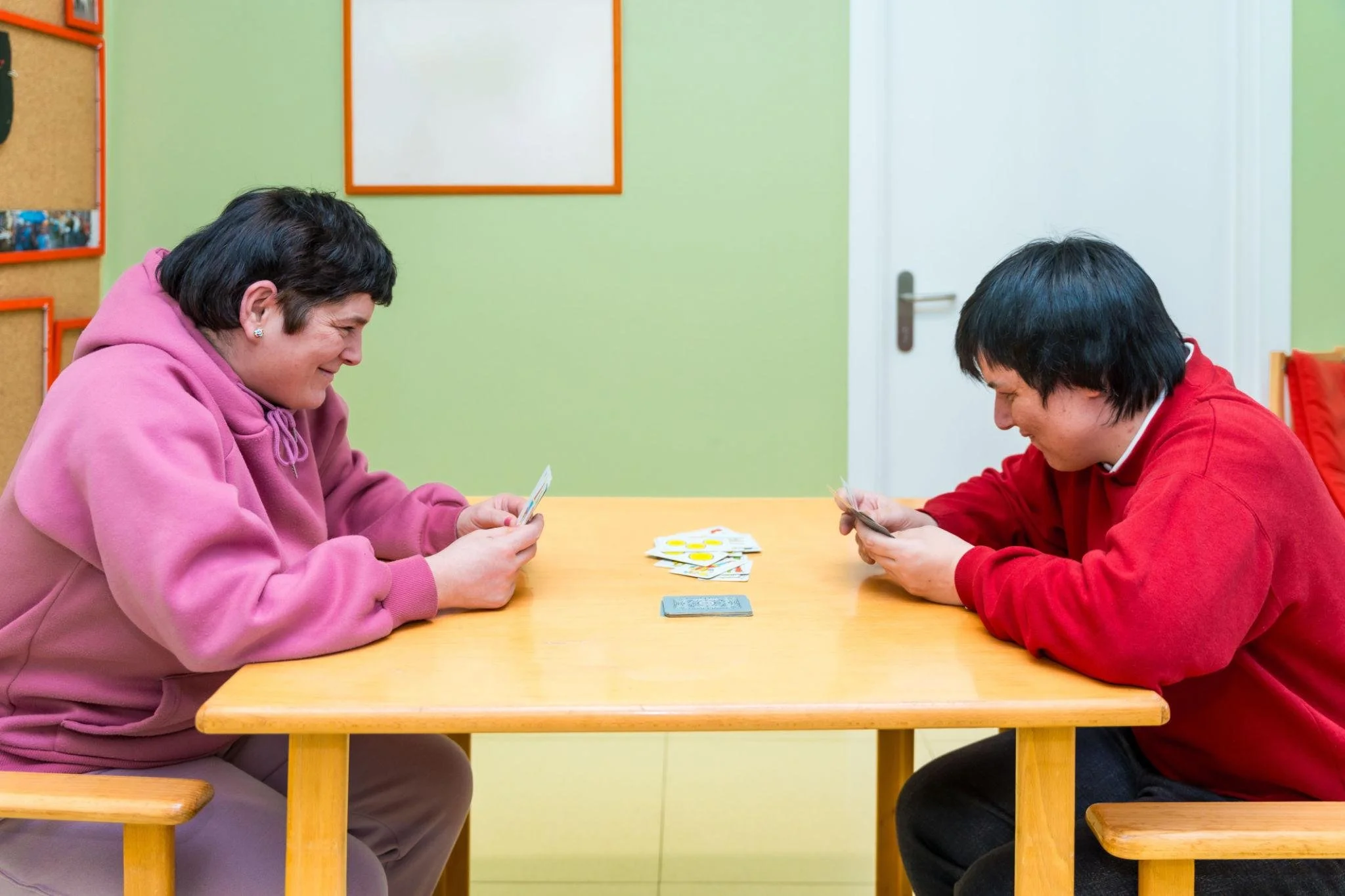 Two women playing a card game at a wooden table in a room with green walls and bulletin boards.