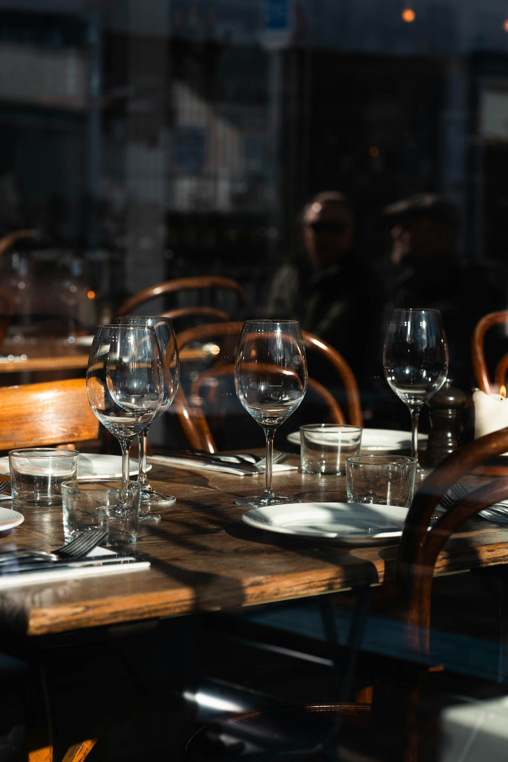 A table set with empty wine glasses, water glasses, plates, and cutlery in a restaurant with large windows and wooden chairs, sunlight casting shadows on the table.