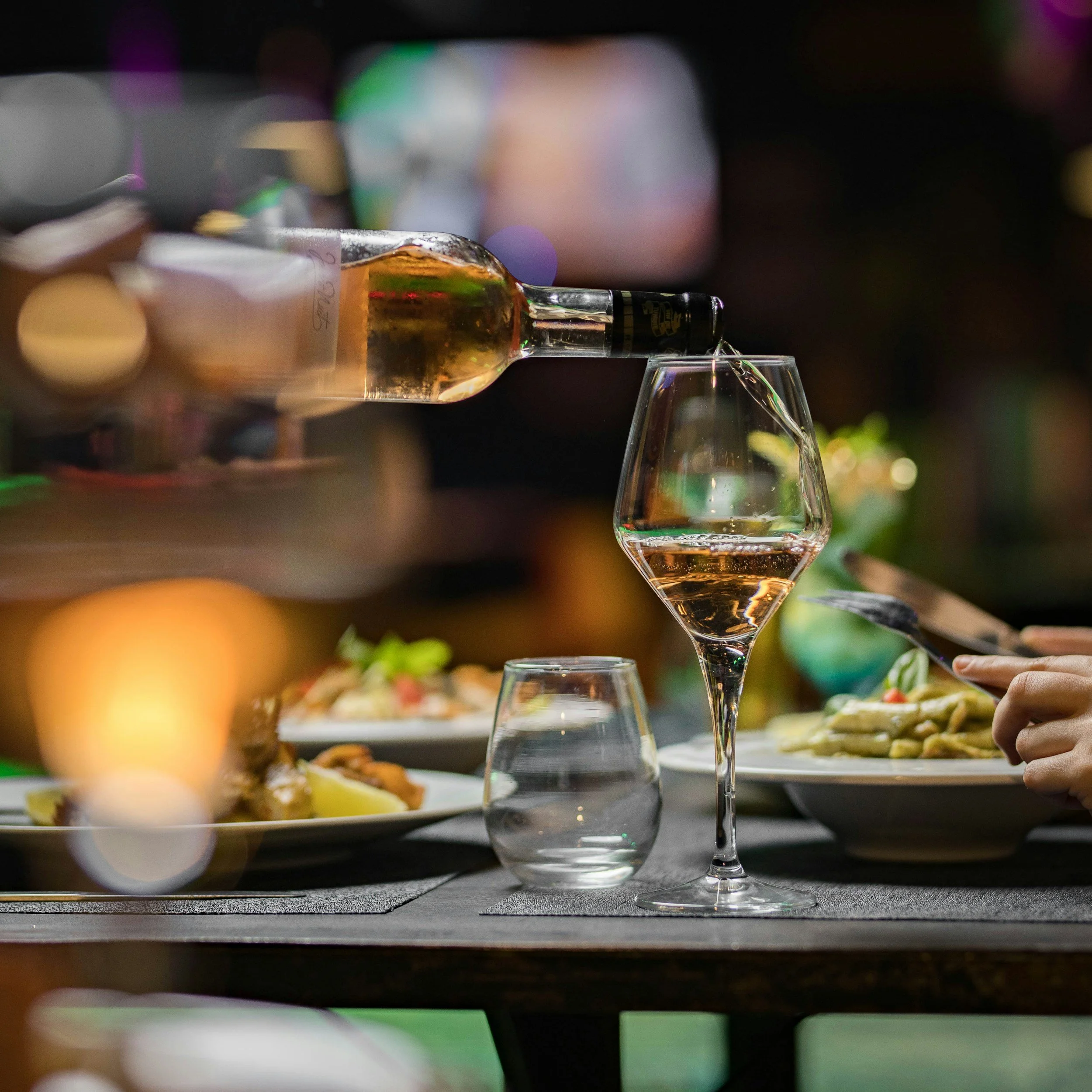 Close-up of a wine glass and a decanter with a blurred background of a dinner setting, including plates, hands, and colorful bokeh lights.